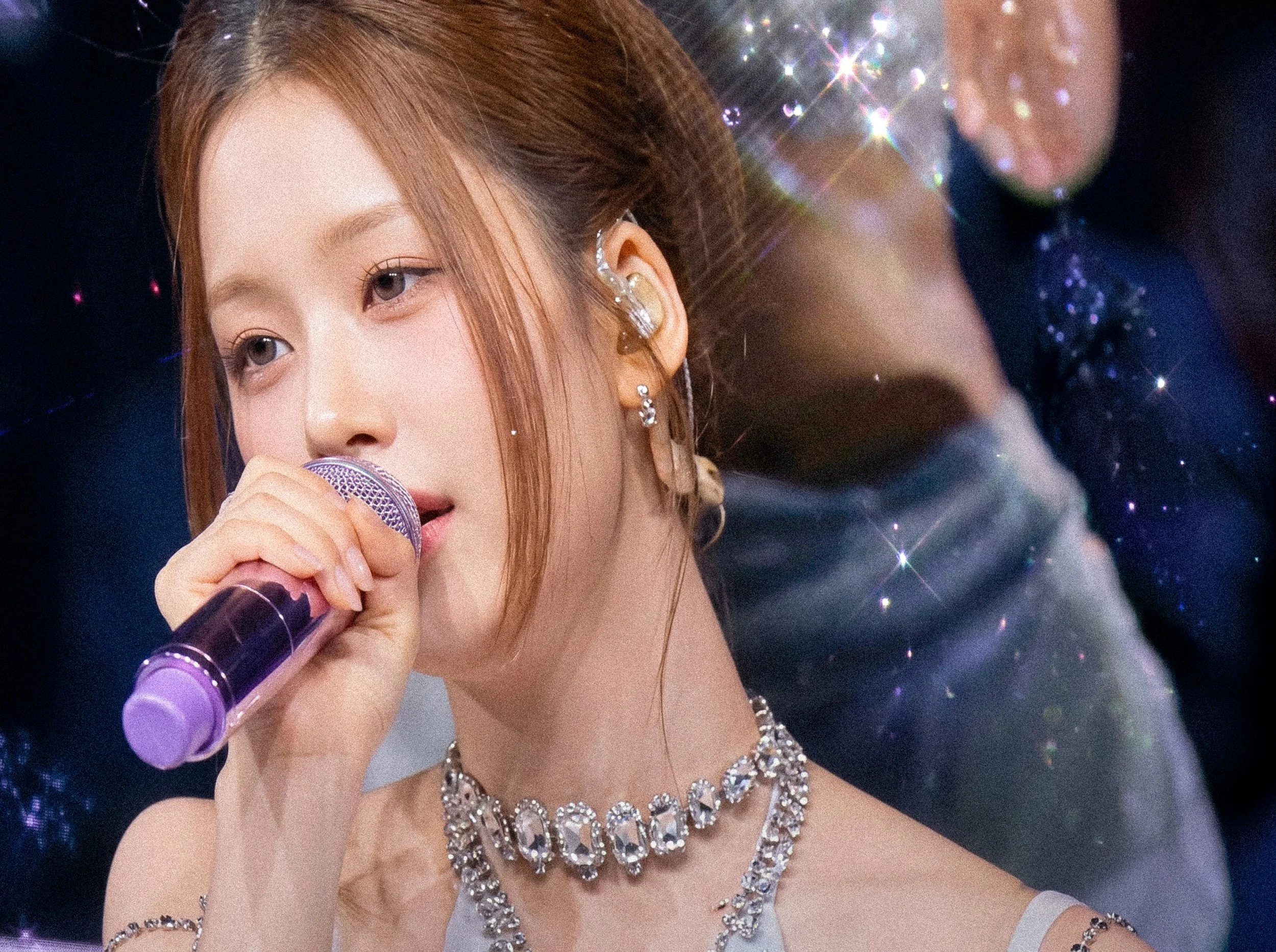 A female performer holding a microphone on stage, wearing jewelry including earrings, a necklace, and a rhinestone headband, with curly hair and a glittering background.