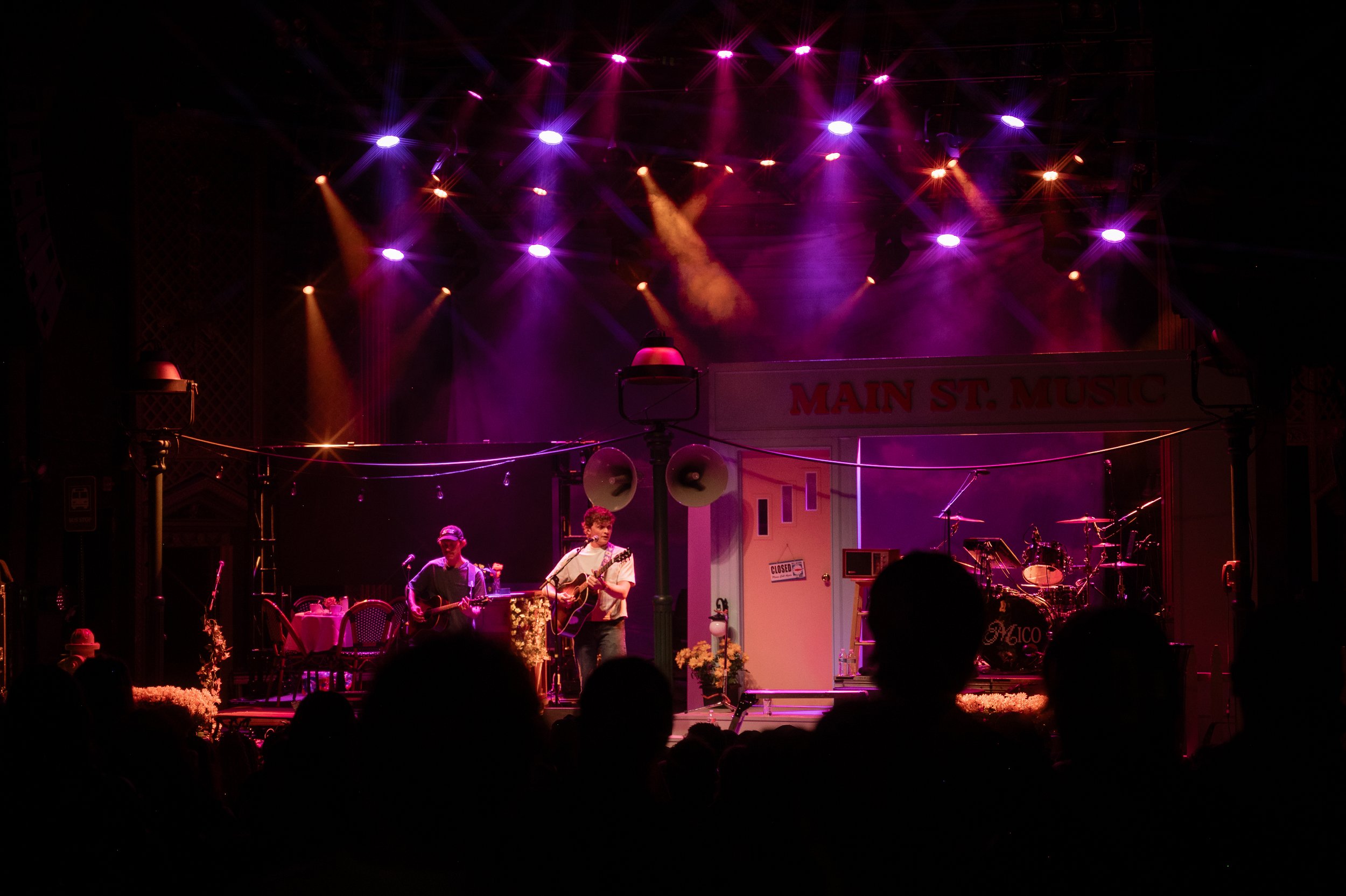 A stage with purple and orange colorful stage lights, a musical band performing with two young boys playing guitars, and audience silhouettes at a music venue named Main St. Music.
