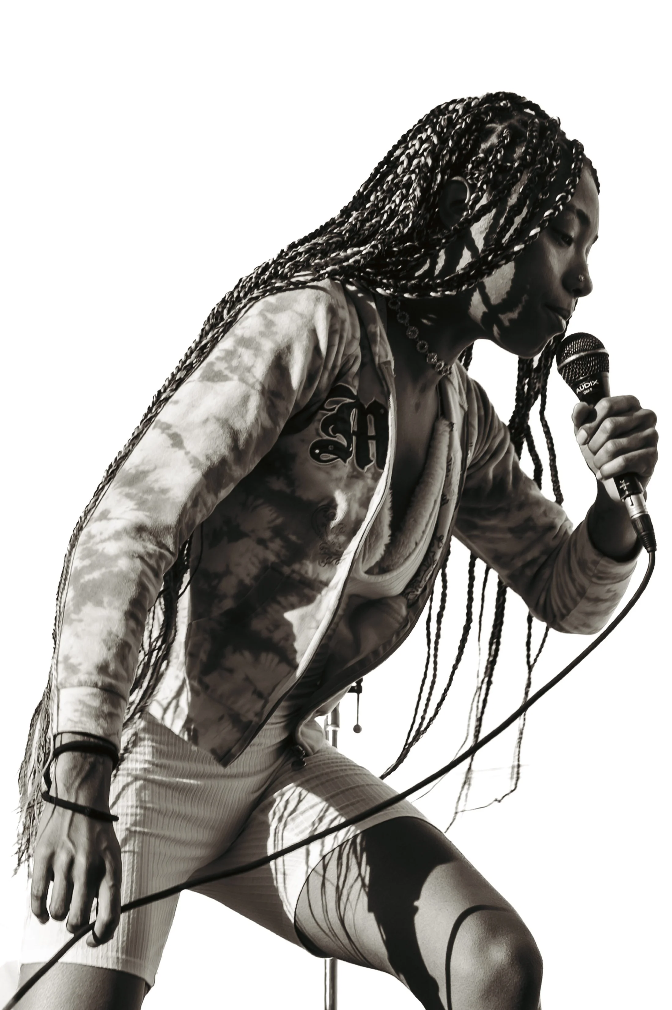 A woman with braided hair performing a dance move, wearing patterned leggings and a jacket, holding a microphone, with shadows cast on her face.