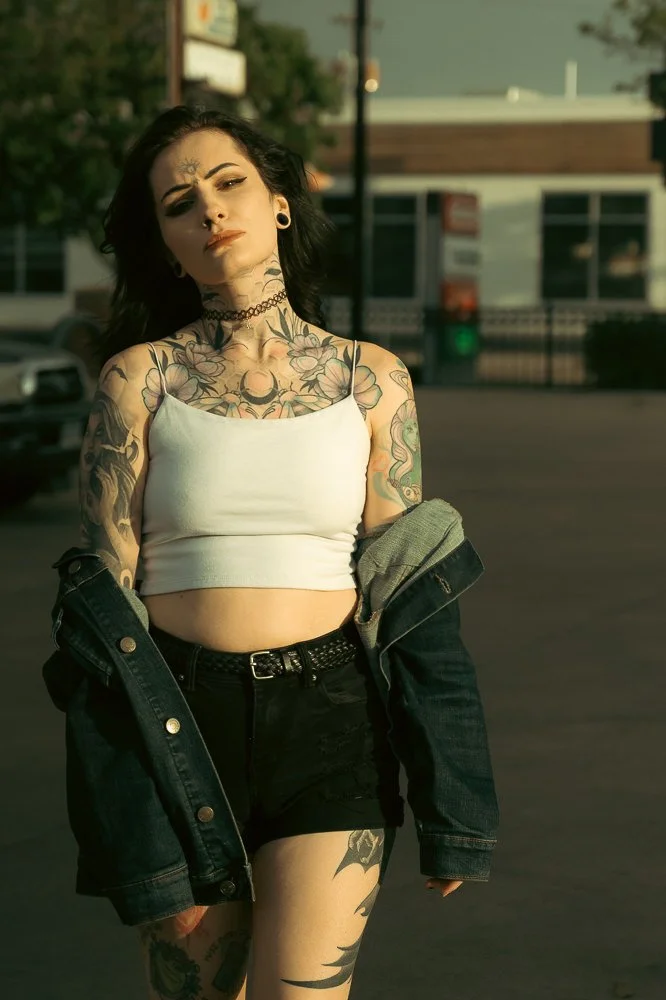 Young woman with tattoos wearing a white crop top, black shorts, and a denim jacket over her shoulders, standing outdoors in a parking lot during late afternoon or early evening.