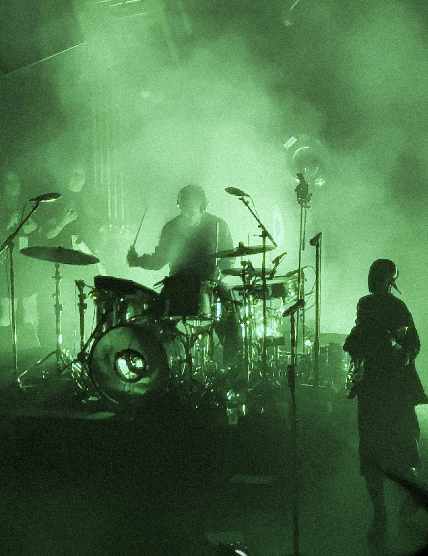 A band performing on stage under green lighting. The drummer is playing drums in the center, with other musicians and equipment visible around him. There are a couple of microphones and a person kneeling or sitting in front of the stage.