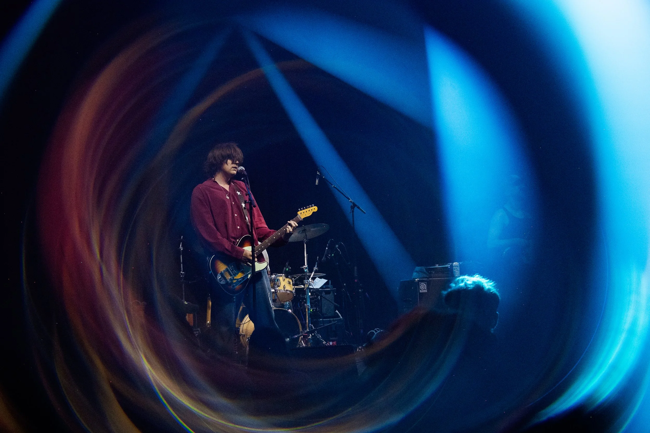A musician playing an electric guitar on stage, framed through a large circular lens with colorful light effects and stage lighting.