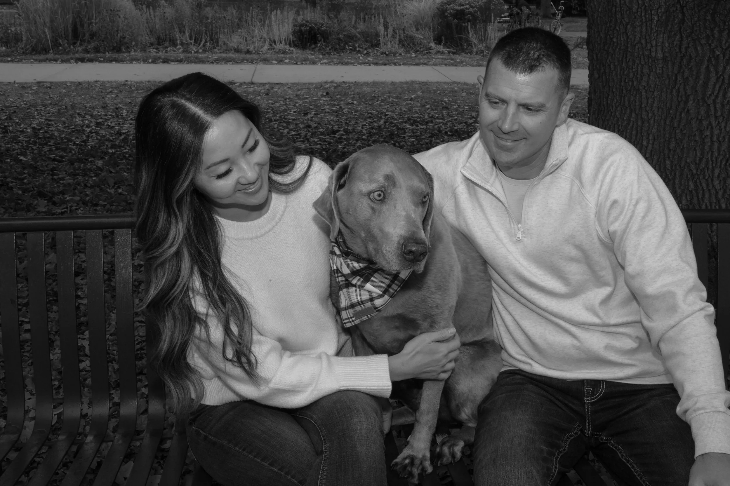 A woman, a man, and a dog sitting on a park bench. The woman is looking down at the dog, and the man is looking at the dog. The dog has striking eyes and is wearing a plaid bandana. The background shows a sidewalk, grass, and trees.