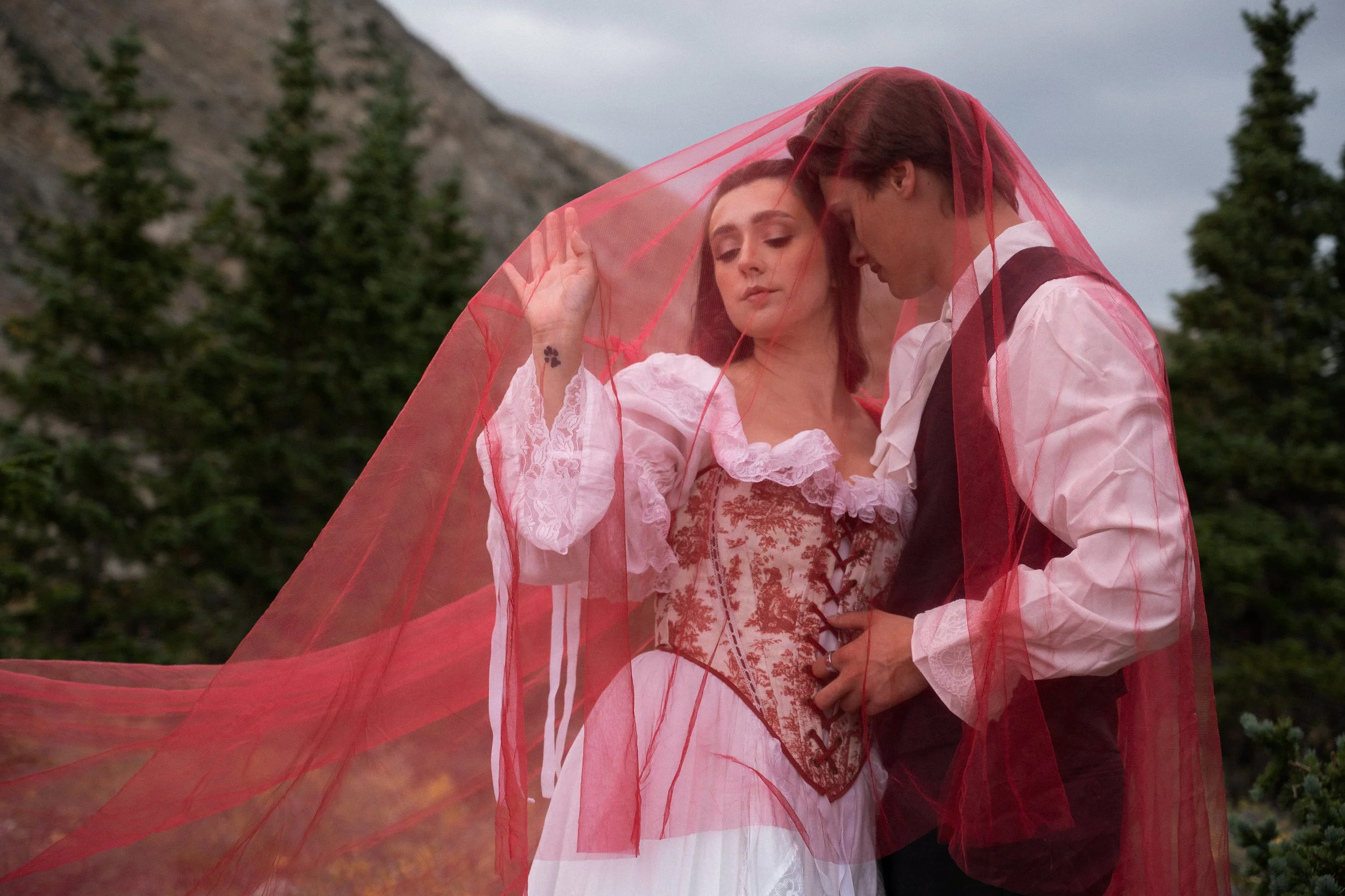 A couple dressed in traditional wedding attire, with the woman in a pink and white gown and the man in a white shirt and black vest, standing close together outdoors under a partly cloudy sky. The woman has a tattoo of a small heart on her wrist and 