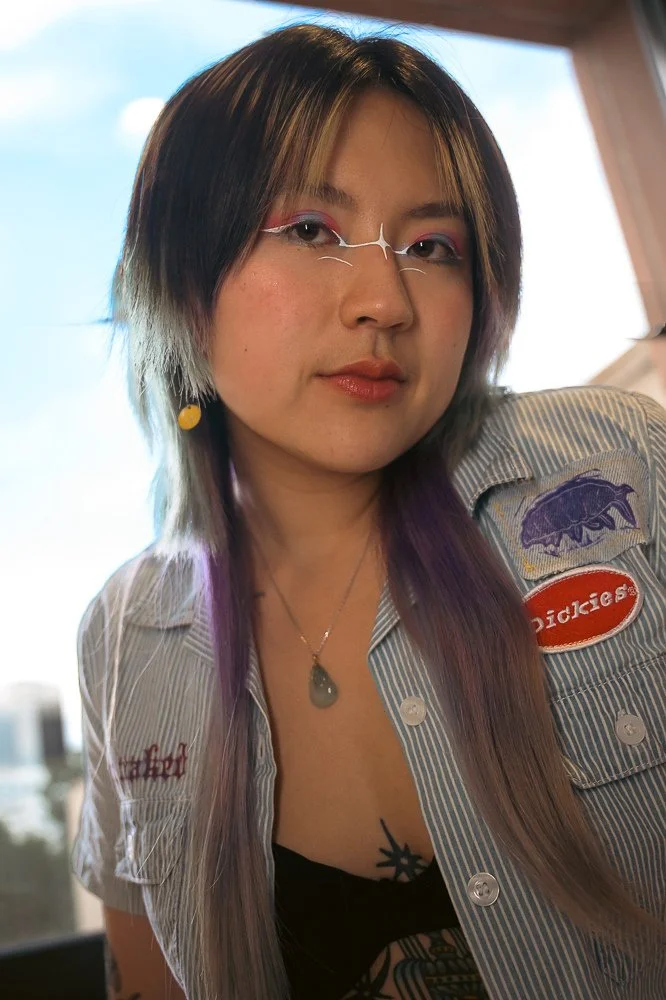 A young woman with multi-colored hair, wearing glasses, a striped shirt with patches, and jewelry, looking into the camera.