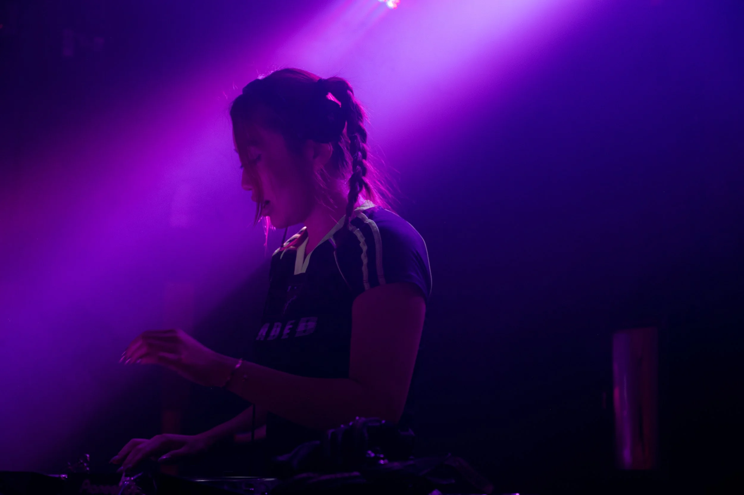 A woman with braided hair, wearing a striped shirt, stands in a dark room illuminated by purple and pink stage lights, possibly performing or DJing.