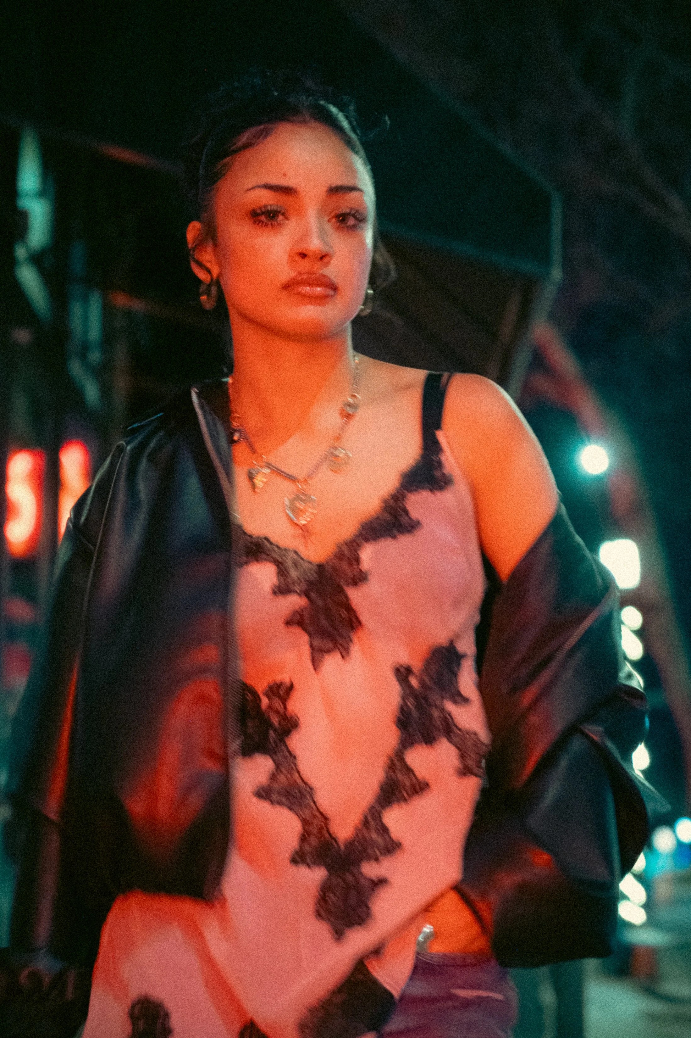 A young woman with dark hair styled with a headband, wearing a lace tank top, with layered necklaces, standing in a dimly lit urban setting with colorful lights.