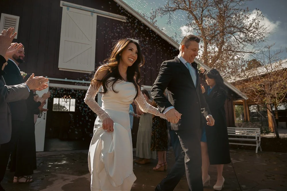 A bride and groom holding hands and walking outdoors at a wedding ceremony, surrounded by guests clapping and celebrating.