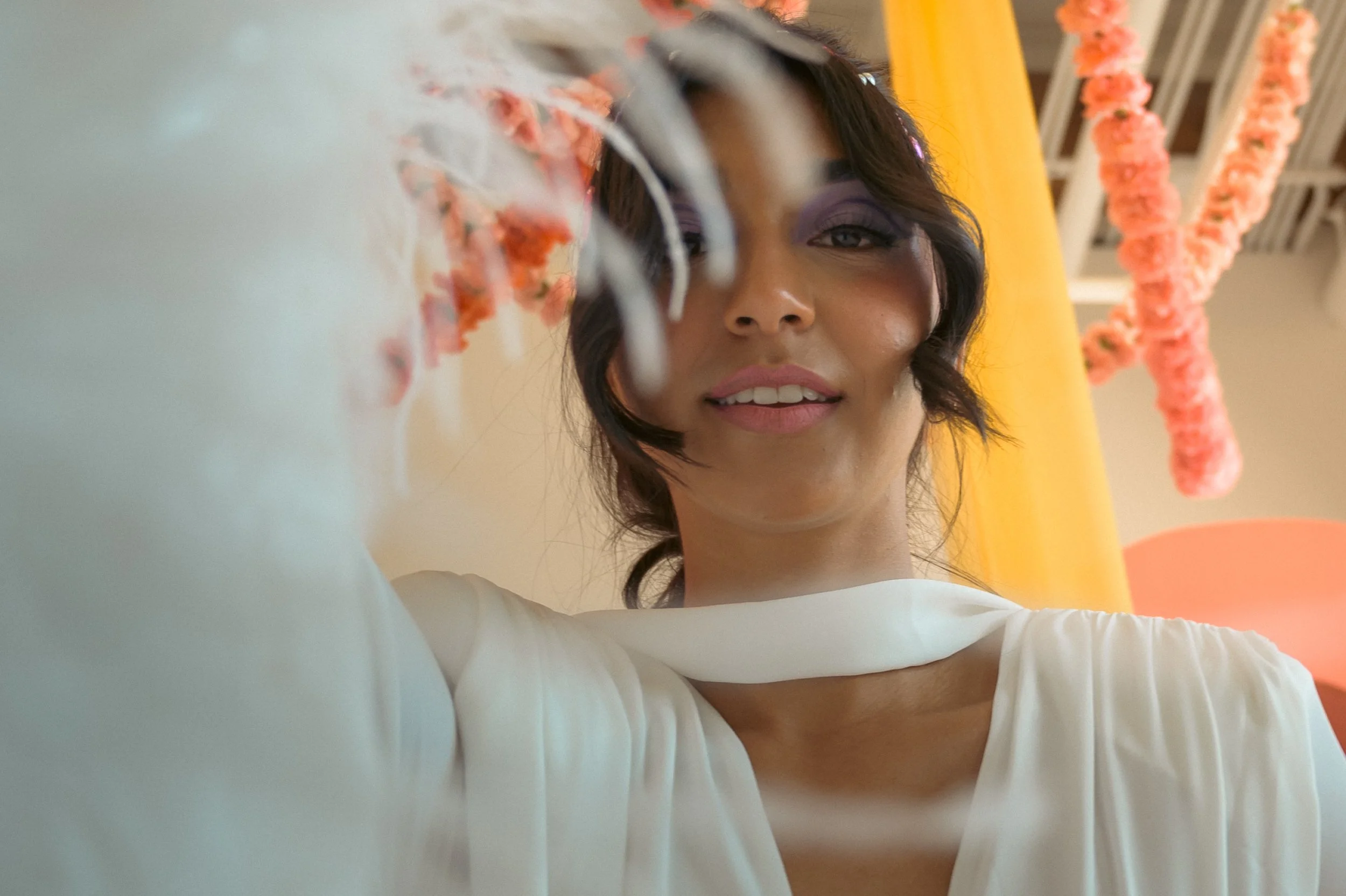 A woman with short dark hair, wearing makeup, is taking a selfie with a focus on her face. She is wearing a white top, and in the background, there is a yellow wall with pink flower garlands hanging.
