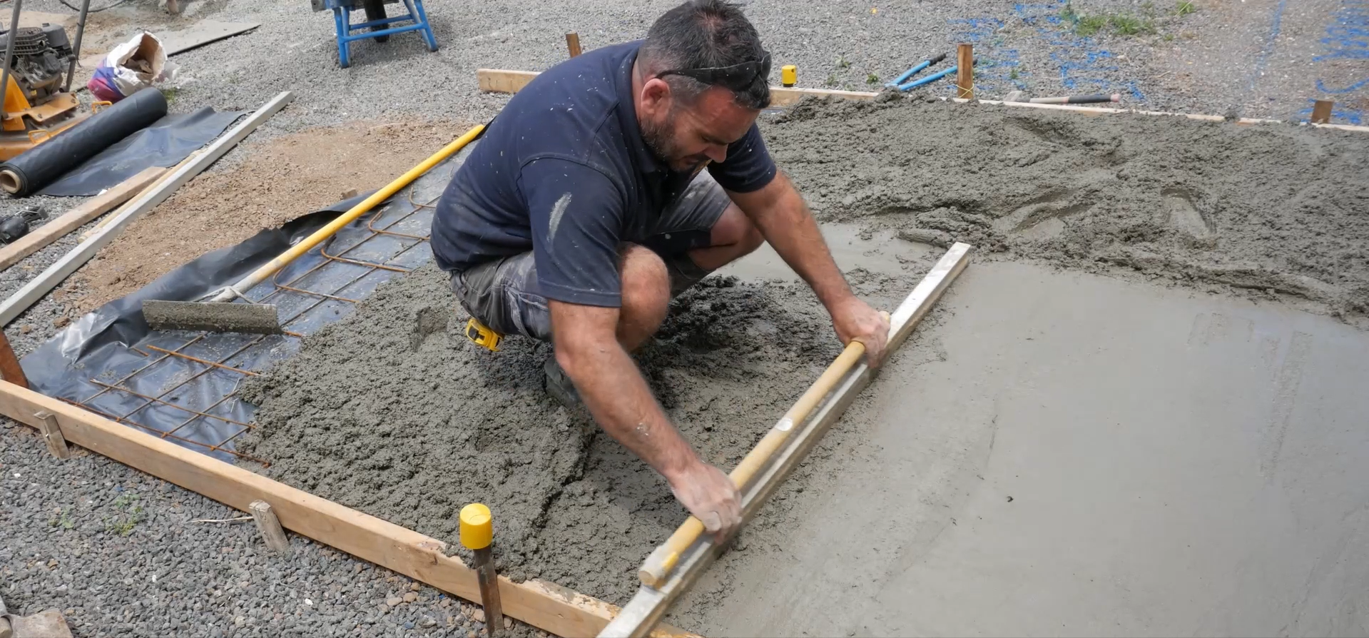 A man working on pouring concrete into a wooden form in a construction site, kneeling and leveling the concrete with a long tool.