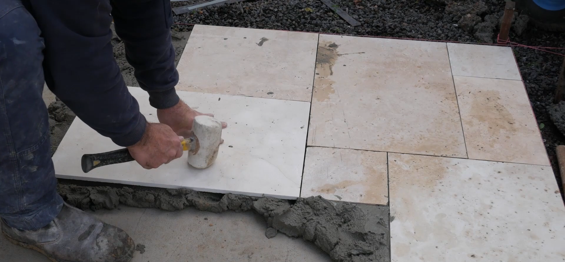 A person installing stone tiles on a floor using a hammer and a rubber mallet, with other tiles already in place and a string line outlining the layout.