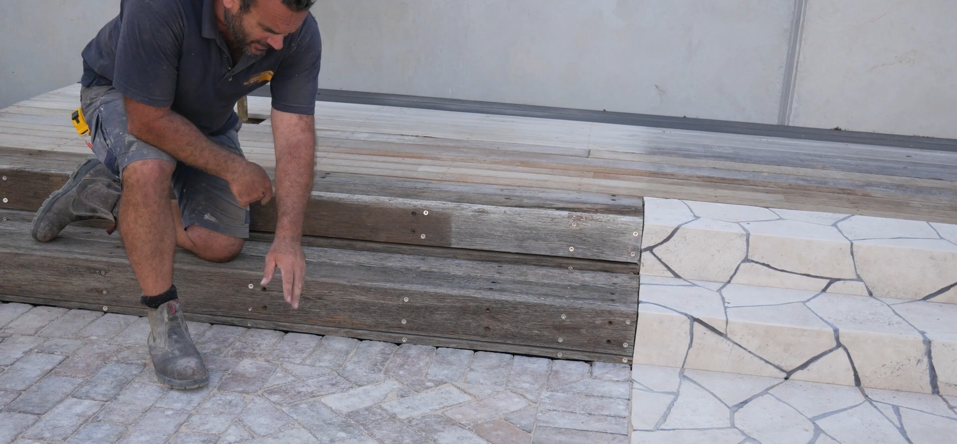 A man kneeling on a wooden deck, working on the edge near stone tiles, with a gray concrete wall in the background.