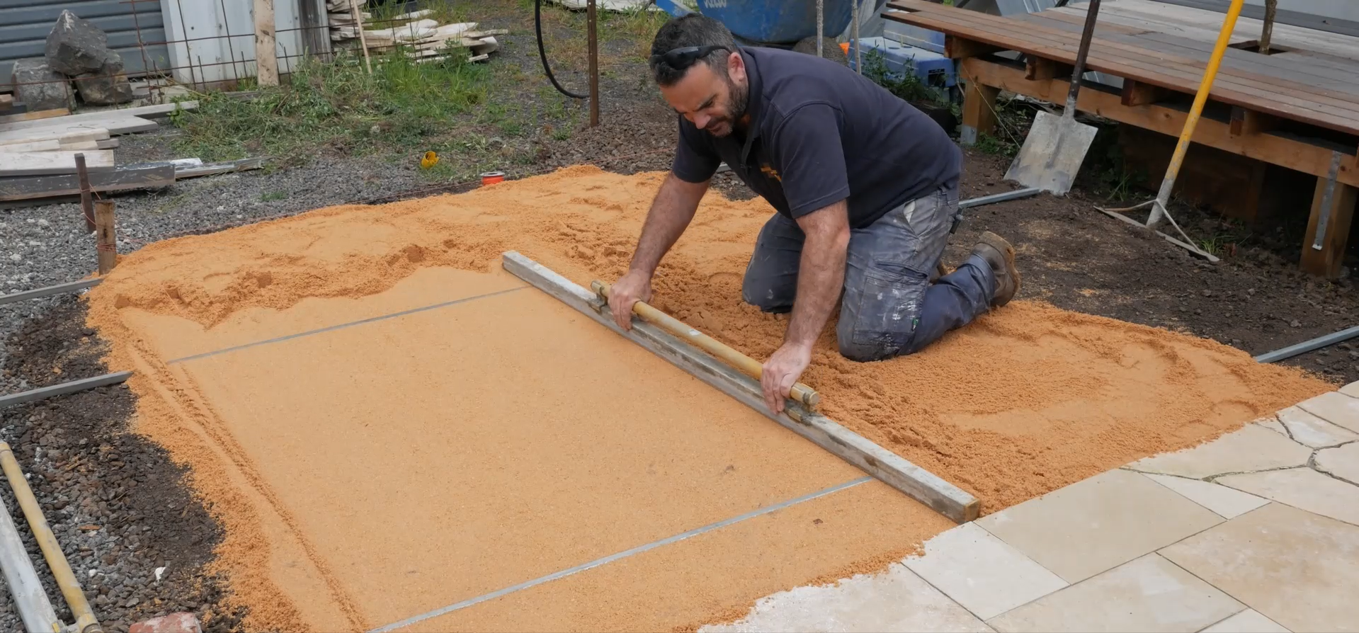 Man working on a construction site, leveling a layer of orange sand or gravel for paving or foundation, using a straight edge and a level.