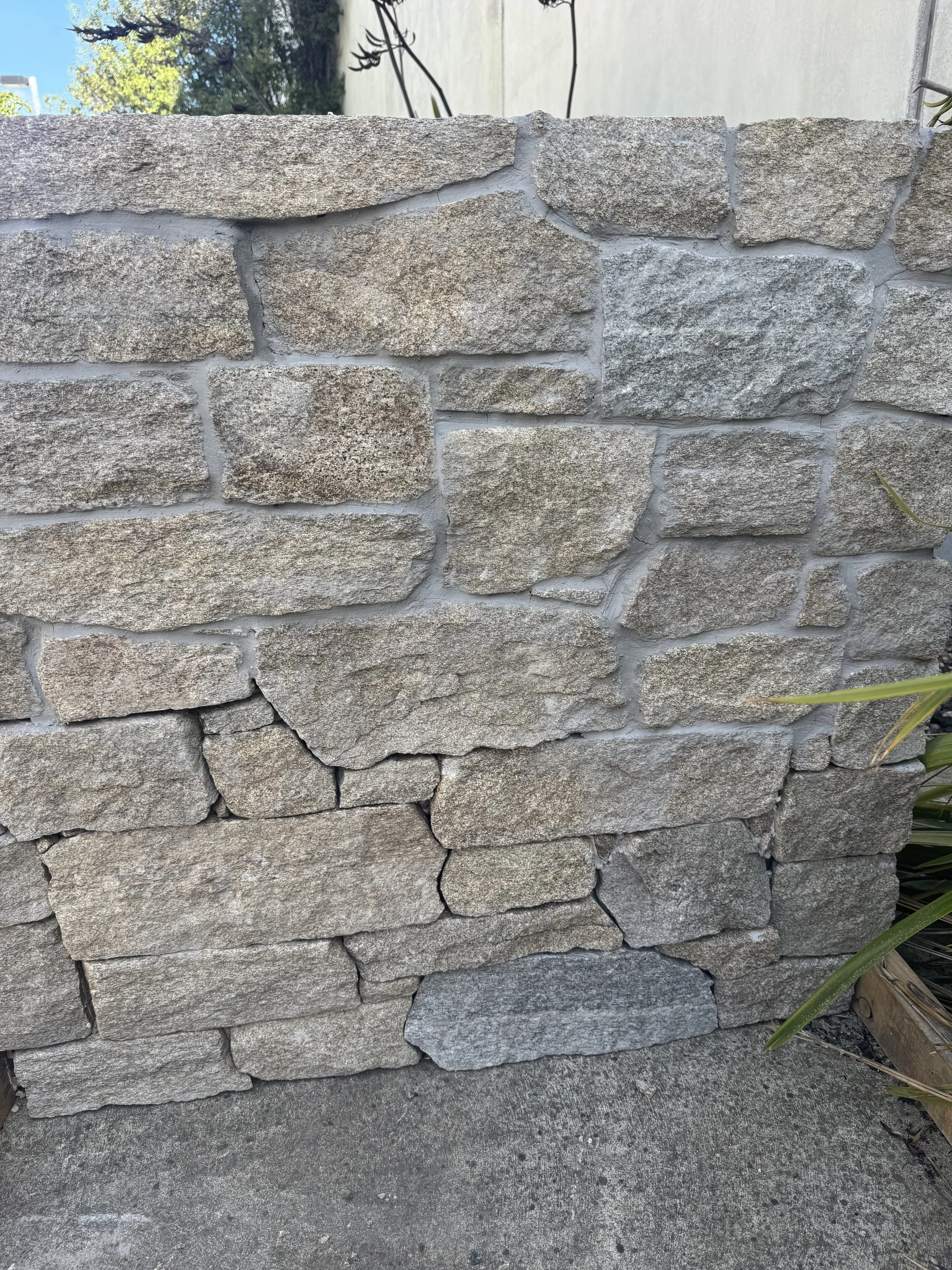 A stone wall with beige and gray stones on a concrete surface, some stones are cracked or broken, and there are plants on the right side.