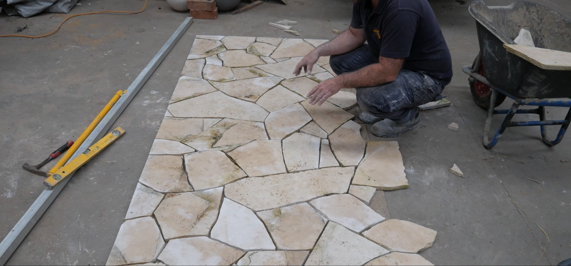 A worker installing irregularly shaped stone tiles on a floor, with tools such as a level, hammer, and wheelbarrow nearby.
