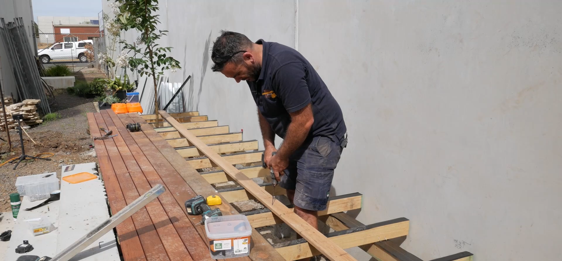 A man working on a woodworking project outdoors, using a drill and working on a wooden frame, with various tools and materials on the table.
