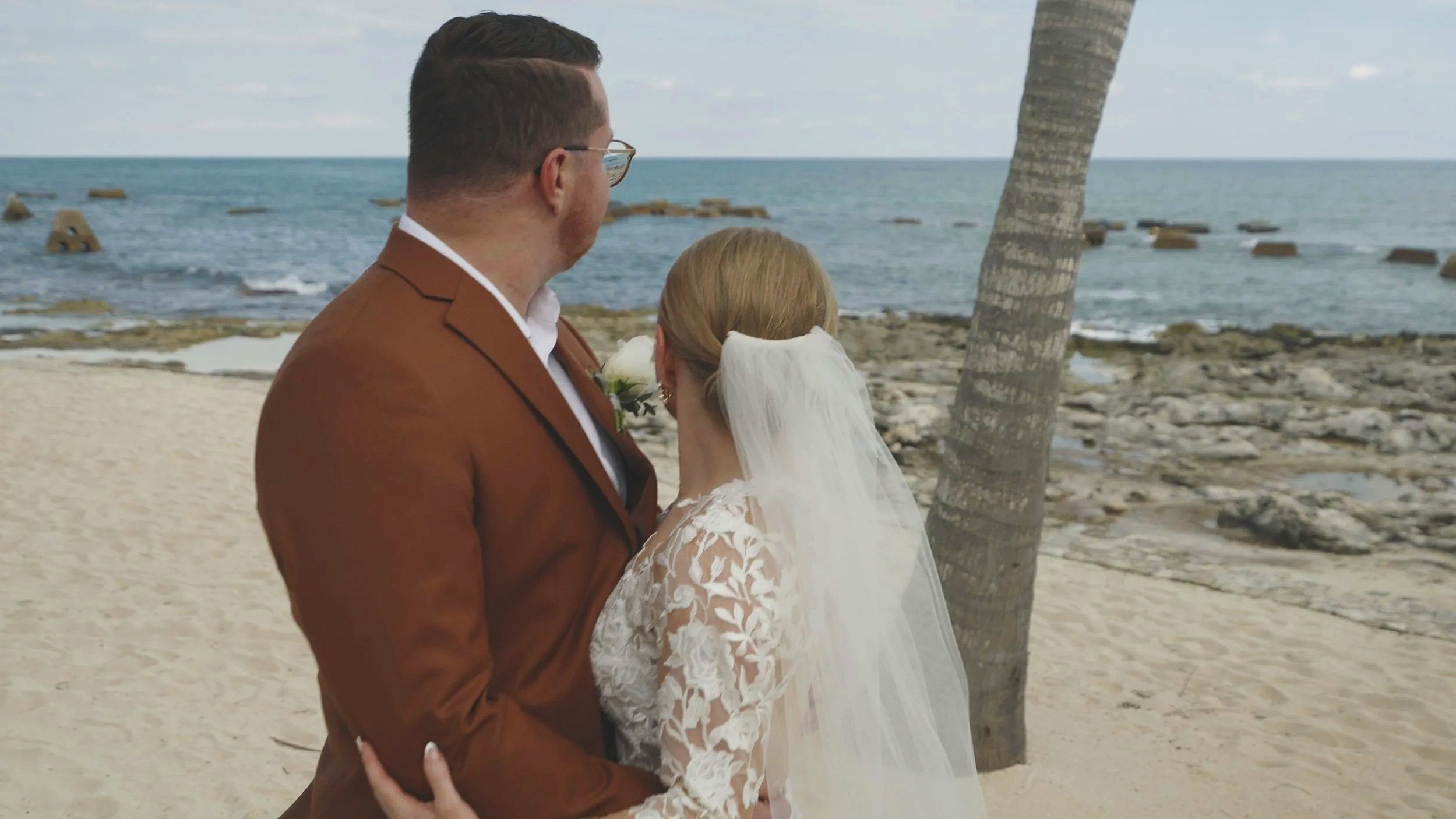 A bride and groom stand close together on a beach, with the ocean behind them and a palm tree to the right.