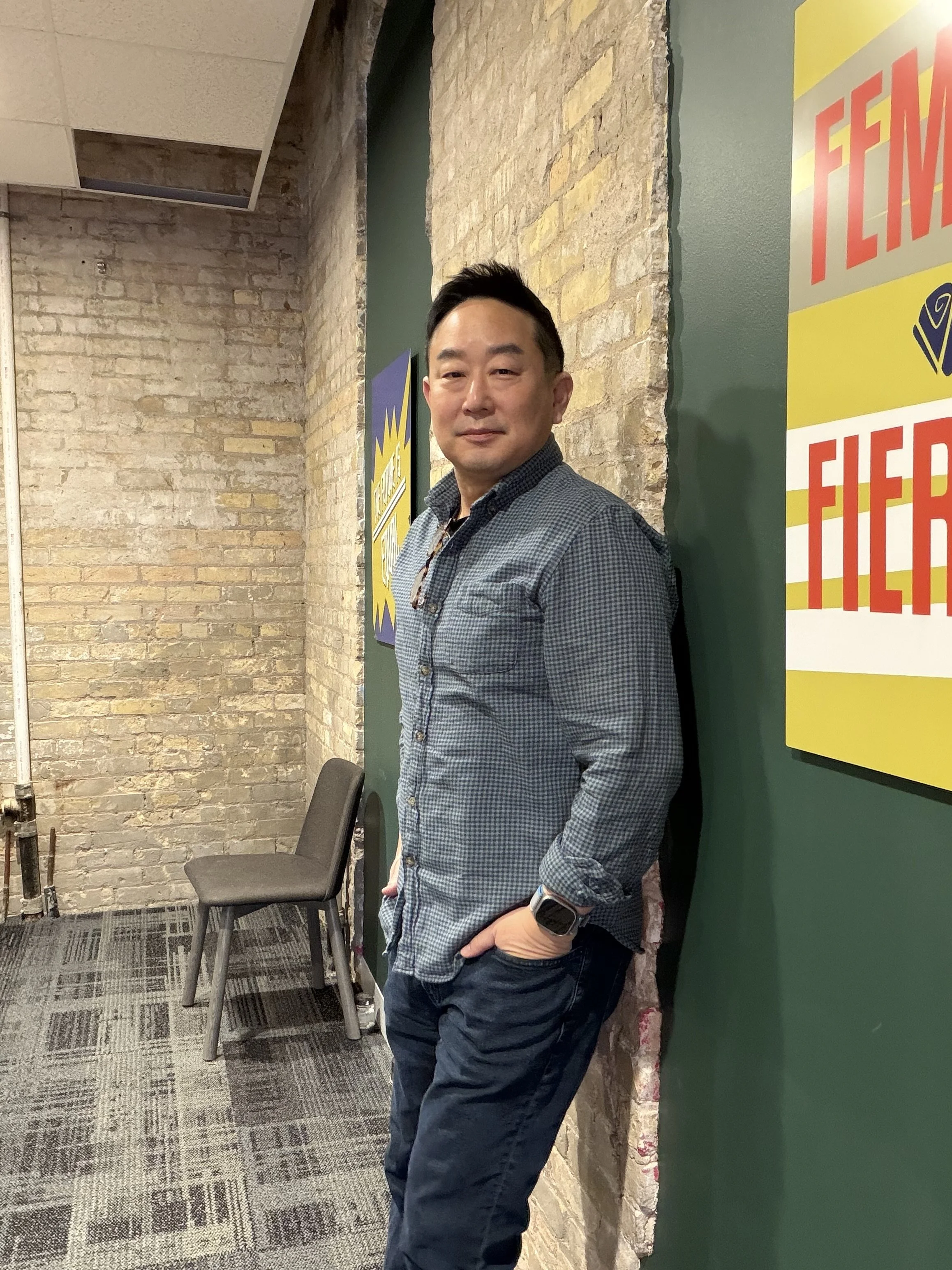 A man with short dark hair and a slight smile, wearing a checkered button-up shirt and dark pants, leaning against a green wall with colorful posters, in an indoor setting with brick and painted walls.