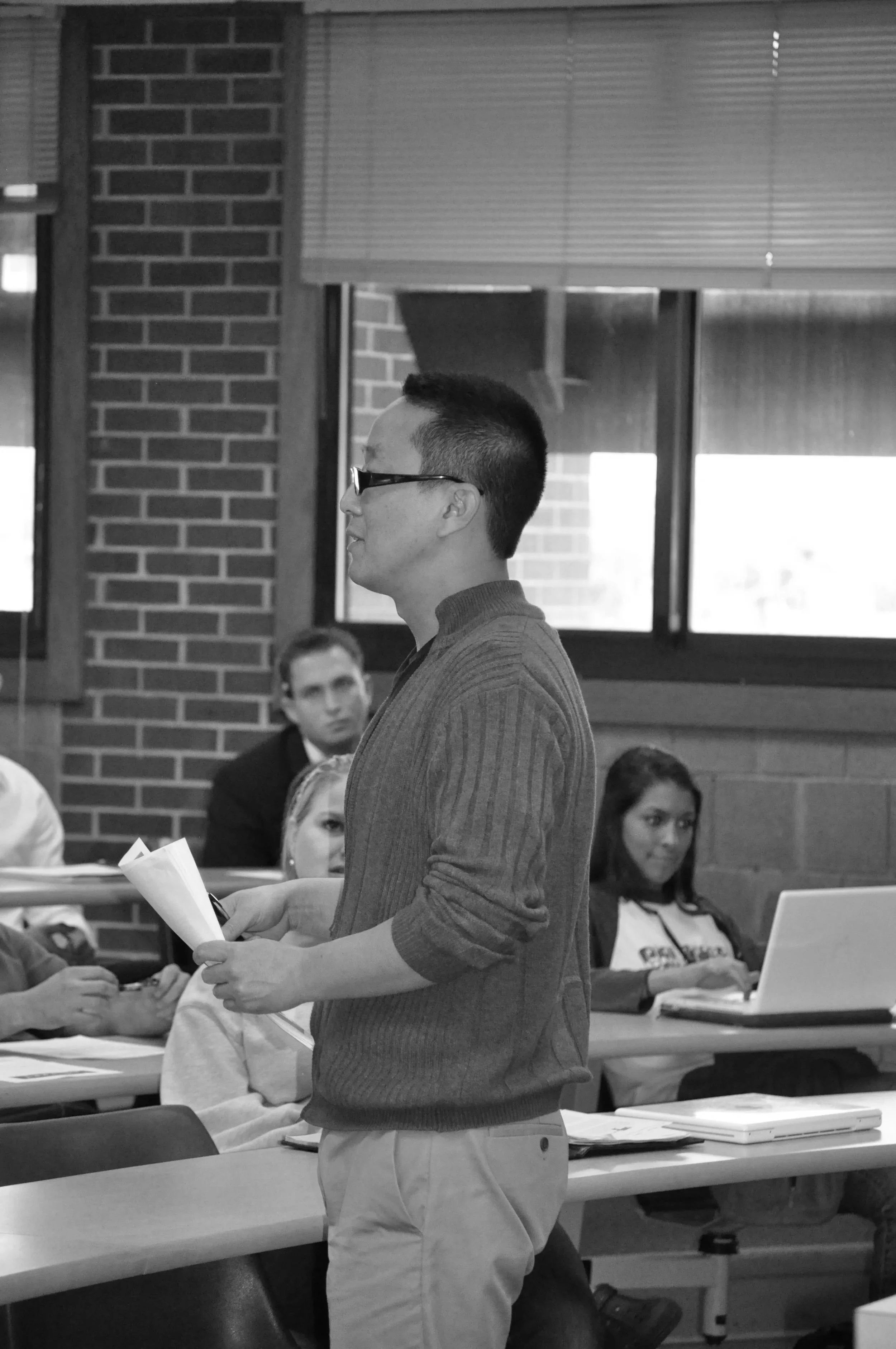 A young man with glasses and a short hairstyle stands in a classroom, holding papers and speaking. Several students are seated at desks, some using laptops or listening, with a brick wall and windows in the background.