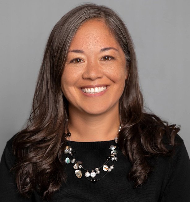 A woman with long brown hair, smiling, wearing a black top and a beaded necklace, against a gray background.