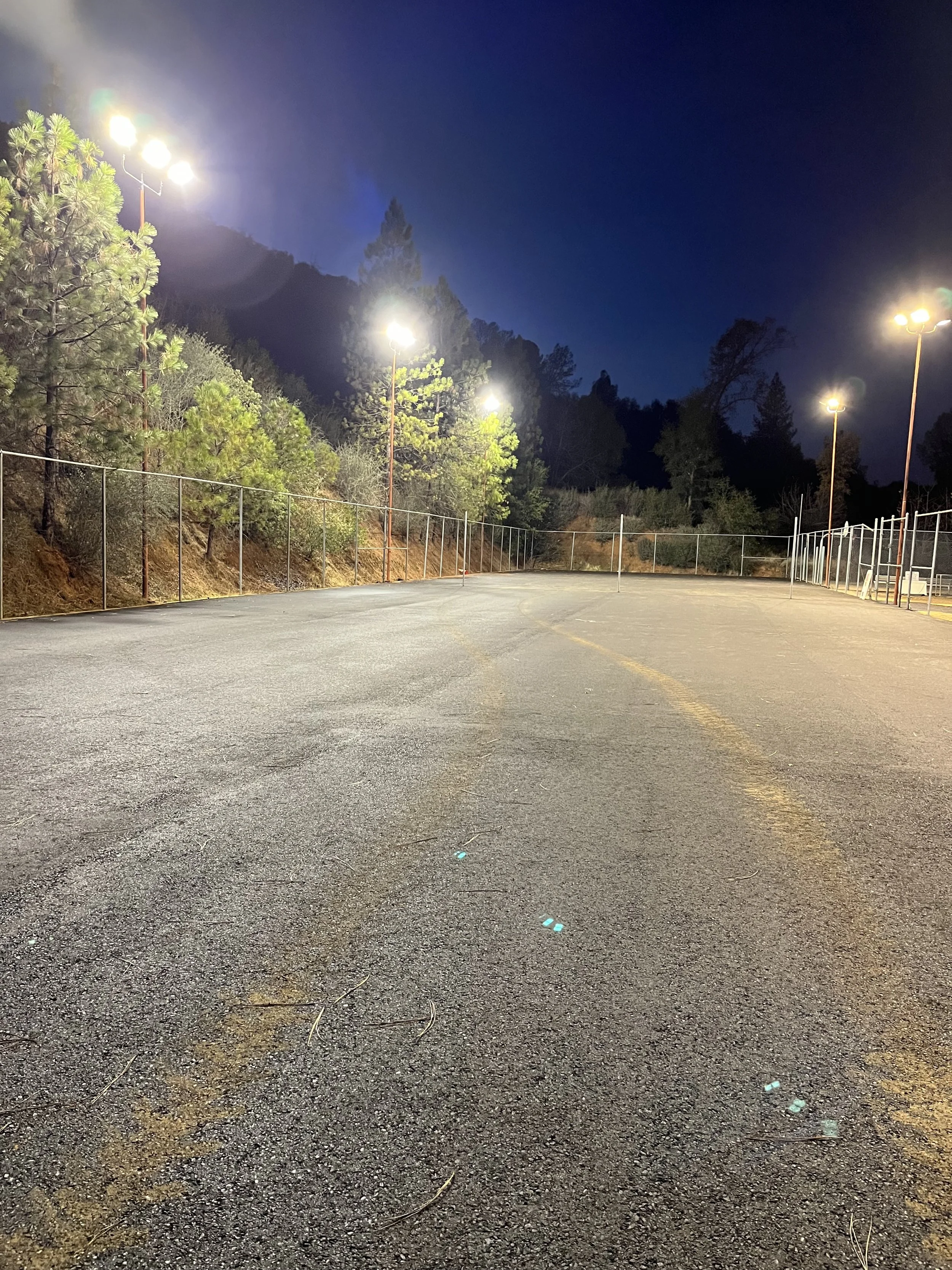 Empty tennis court at night with fence, tall floodlights, surrounded by trees, and a dark sky.