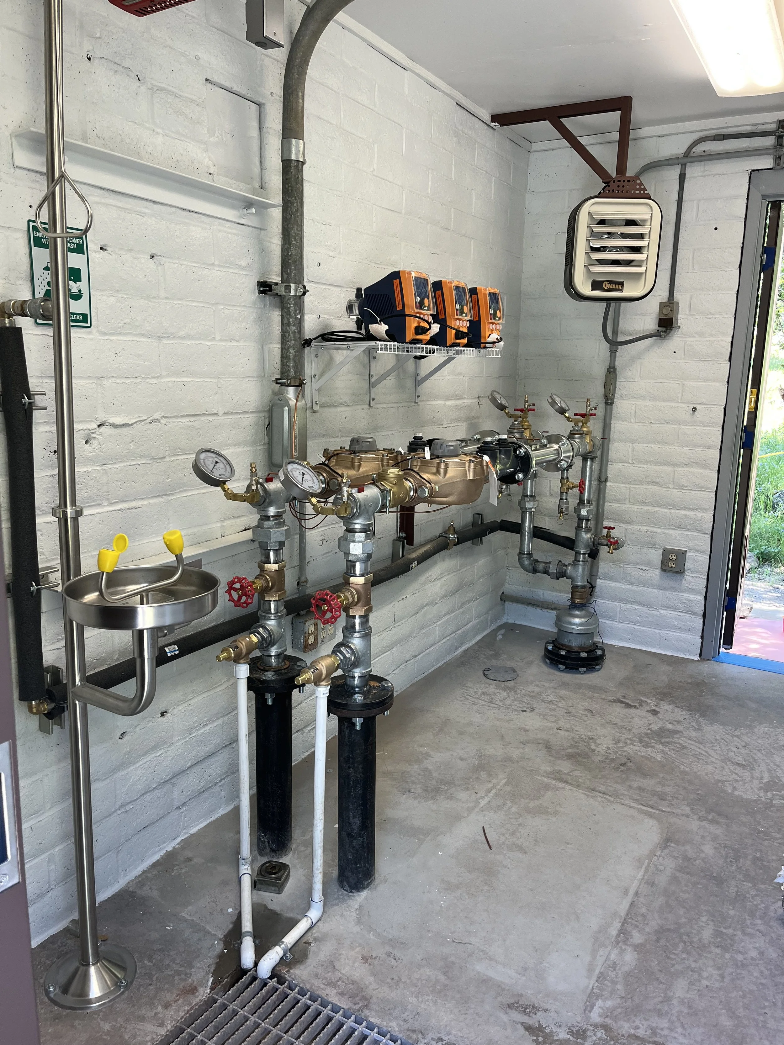 Mechanical room with pipes, gauges, and meters, containing a wall-mounted air conditioning unit, shelf with orange and black devices, and an open door leading outside.