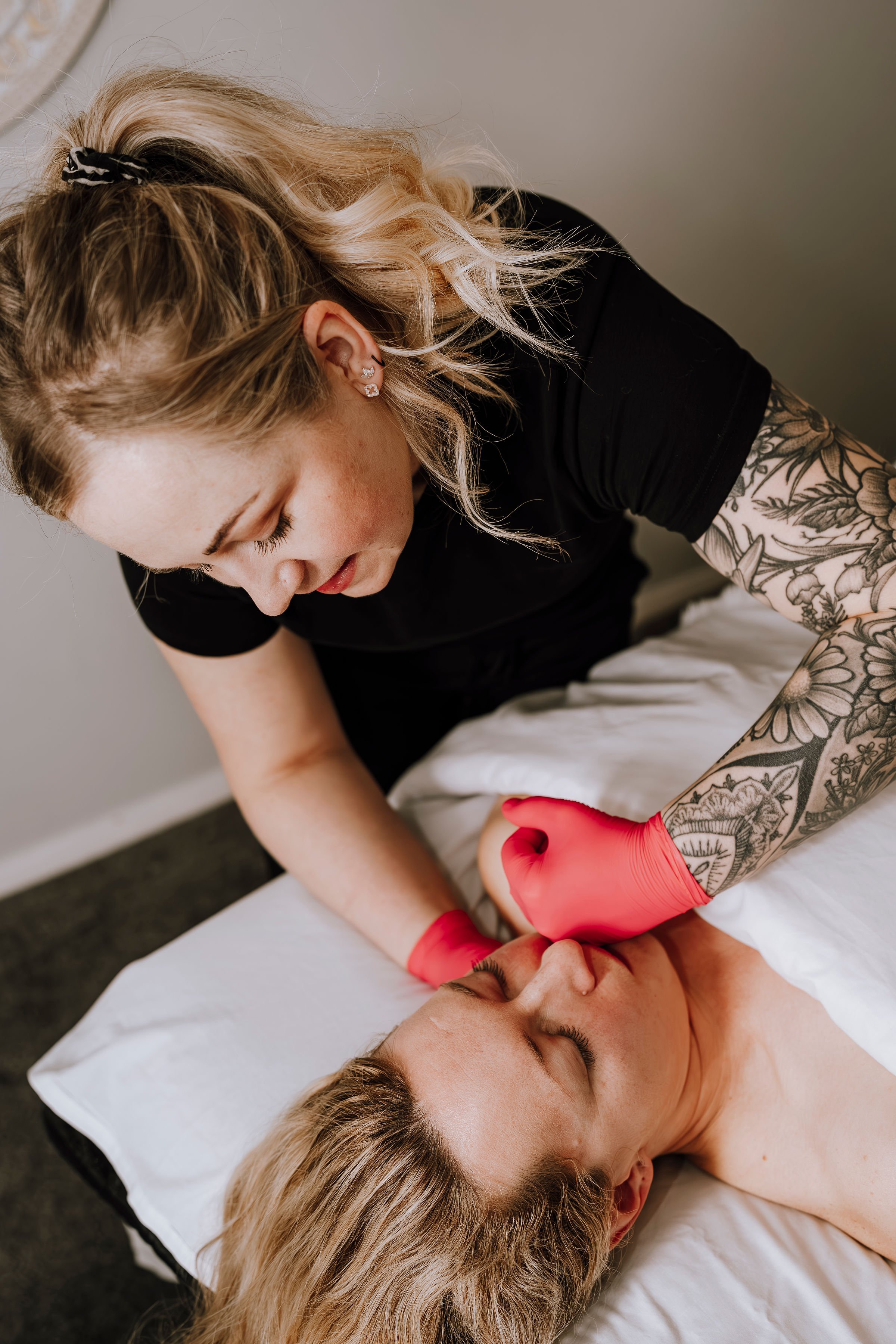 A woman with blonde hair and tattoos on her arm is performing a medical procedure on a woman lying on a bed, using red medical gloves.