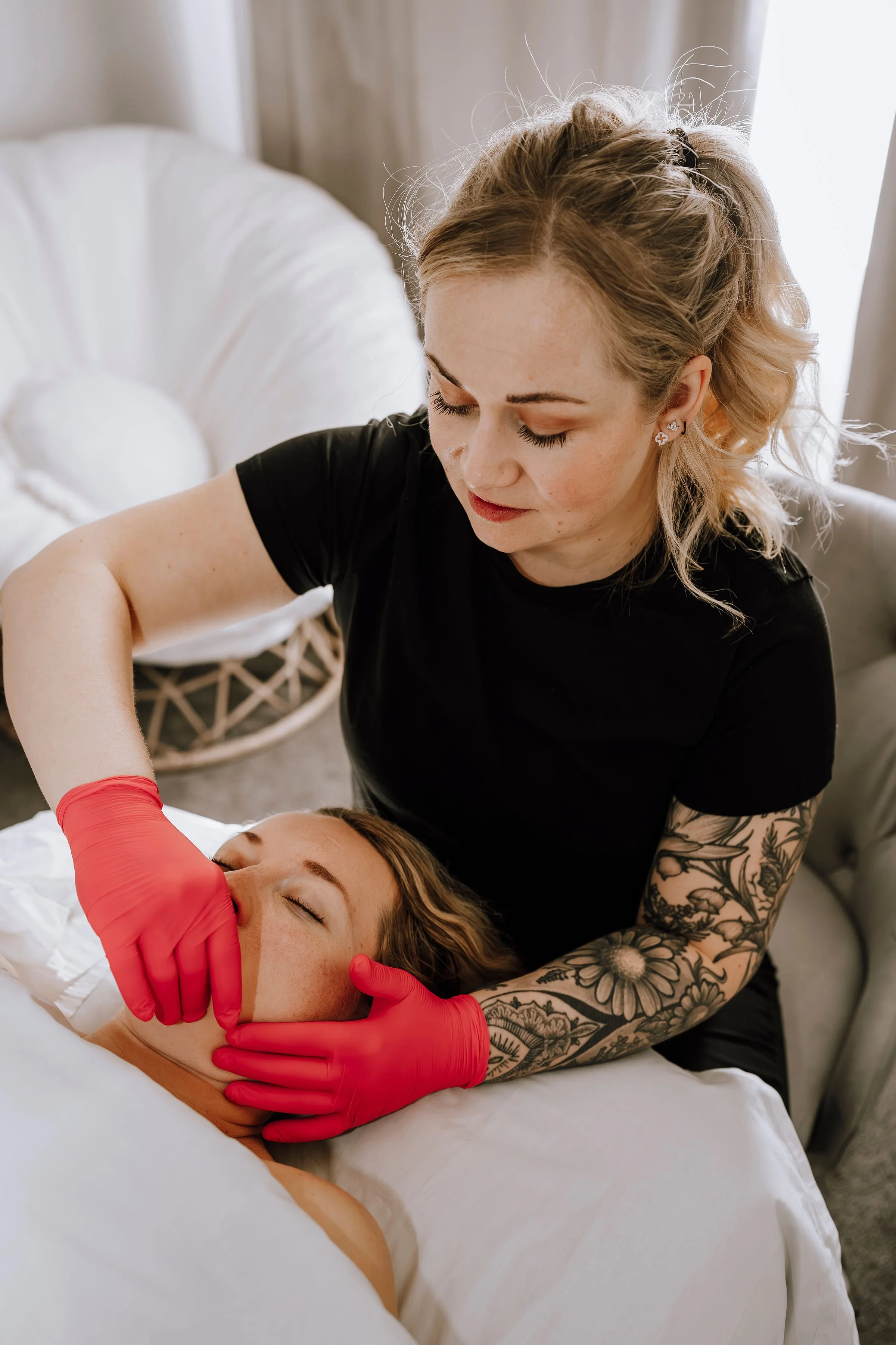 A woman with blonde curly hair, wearing a black shirt and red gloves, providing a facial treatment to another woman with brown hair lying on a bed, in a cozy room with soft lighting.