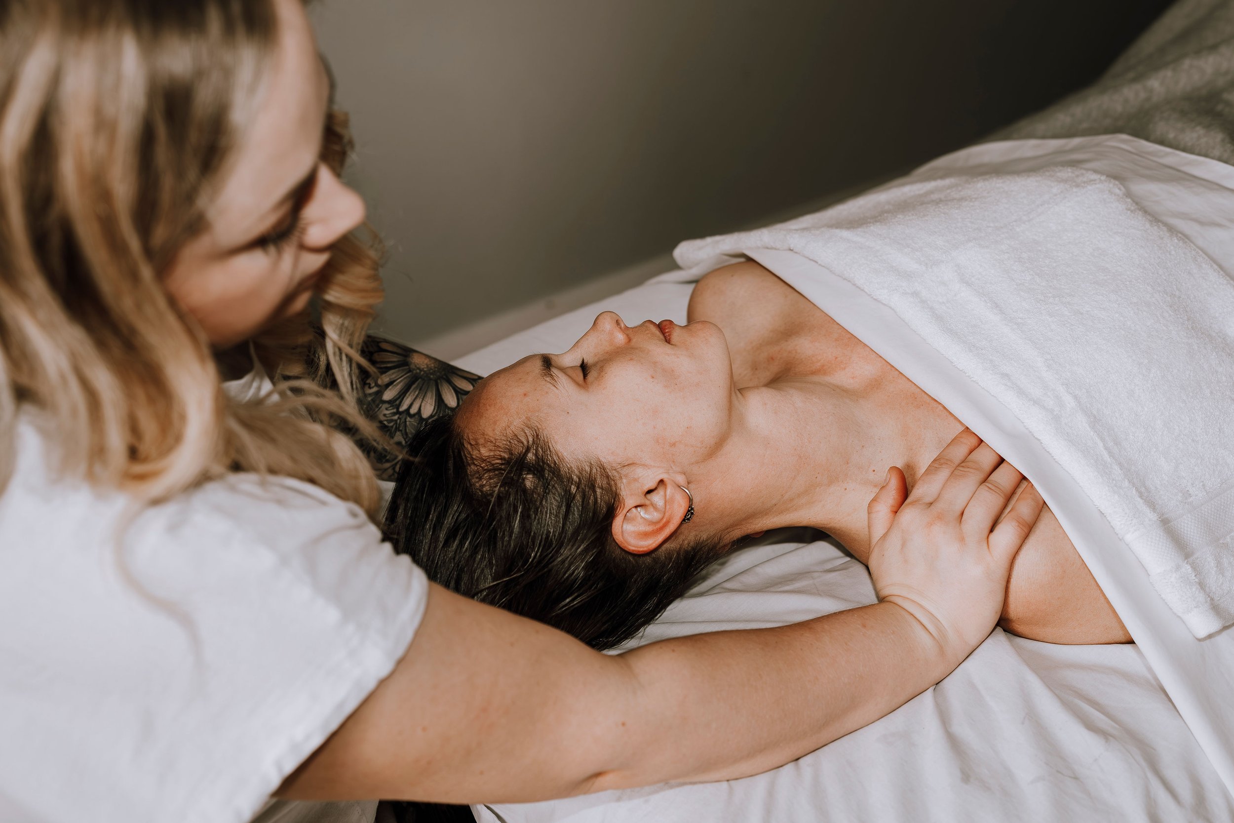 Woman lying on a bed with a towel, smiling at another woman who is leaning over her, touching her shoulder.