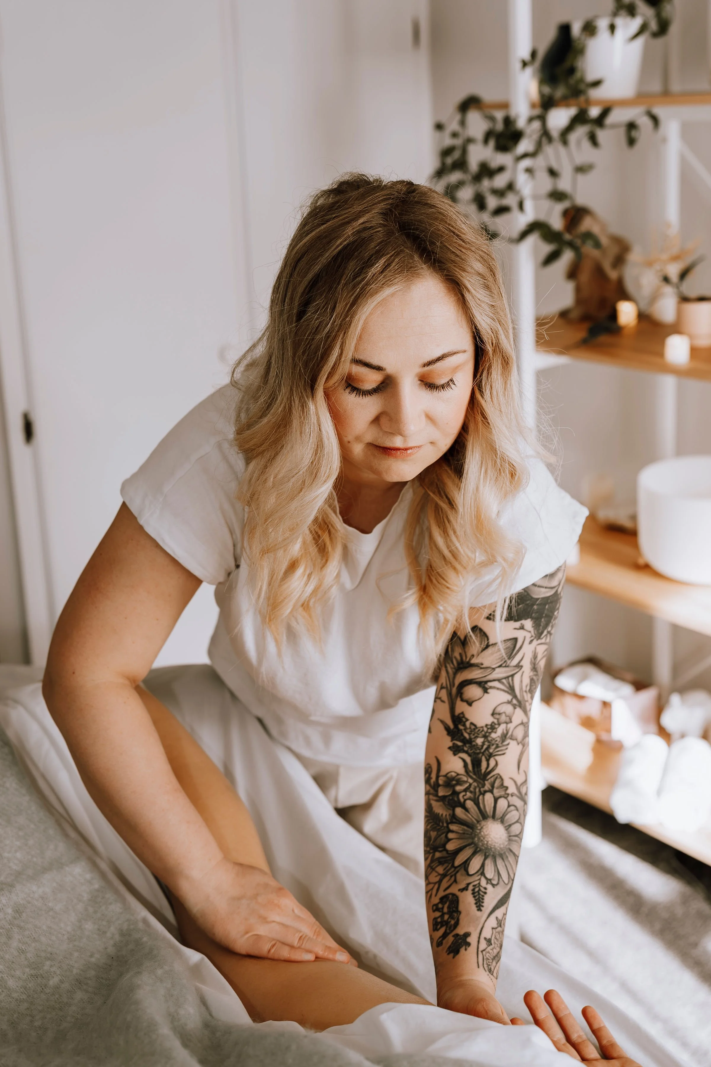 A woman with long, wavy blonde hair giving a leg massage, sitting on a bed in a room with wooden shelves holding candles, plants, and wellness items.