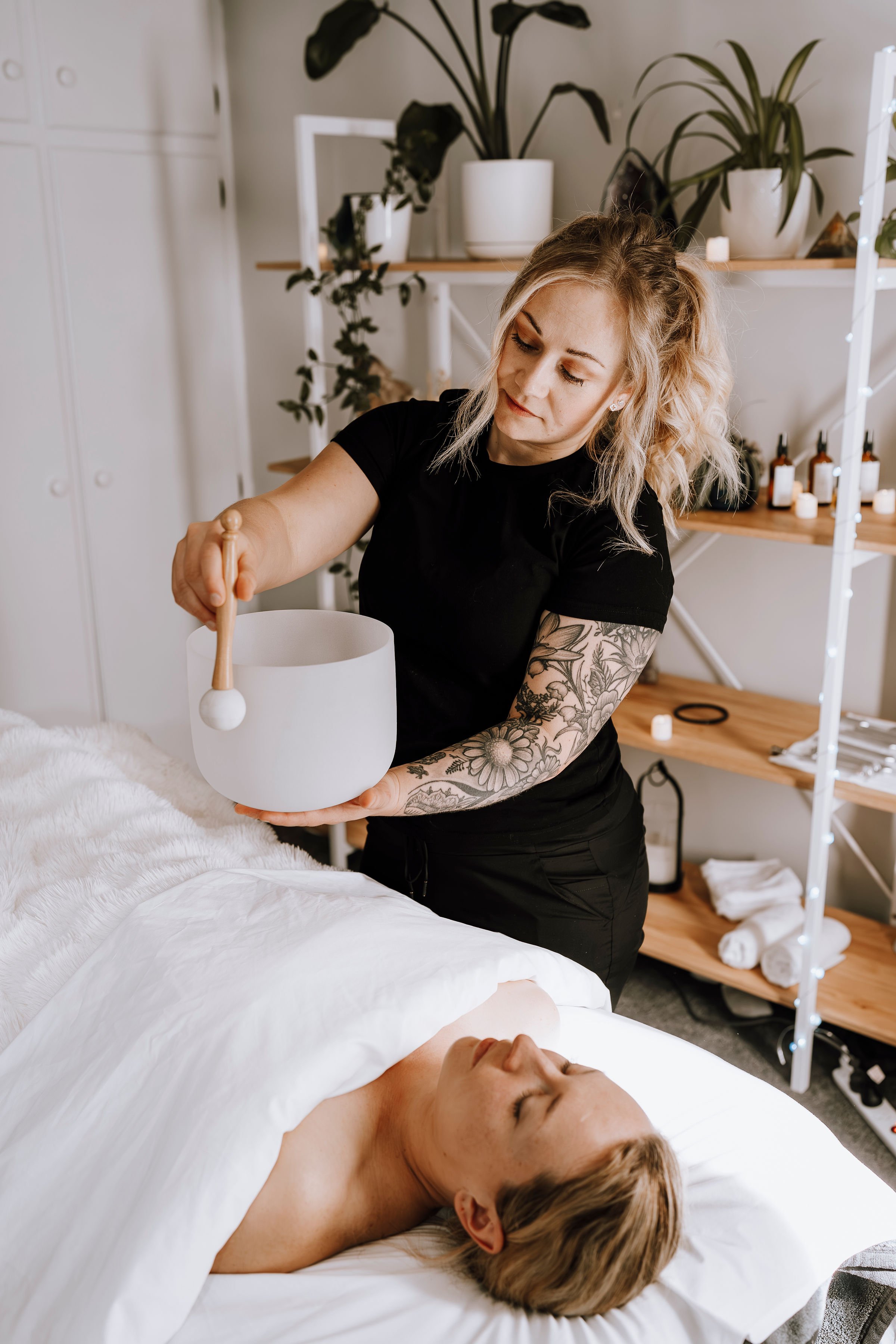 A woman with tattoos on her arm performs sound therapy with a singing bowl on a bed in a wellness room, while a person lies peacefully under a white blanket.