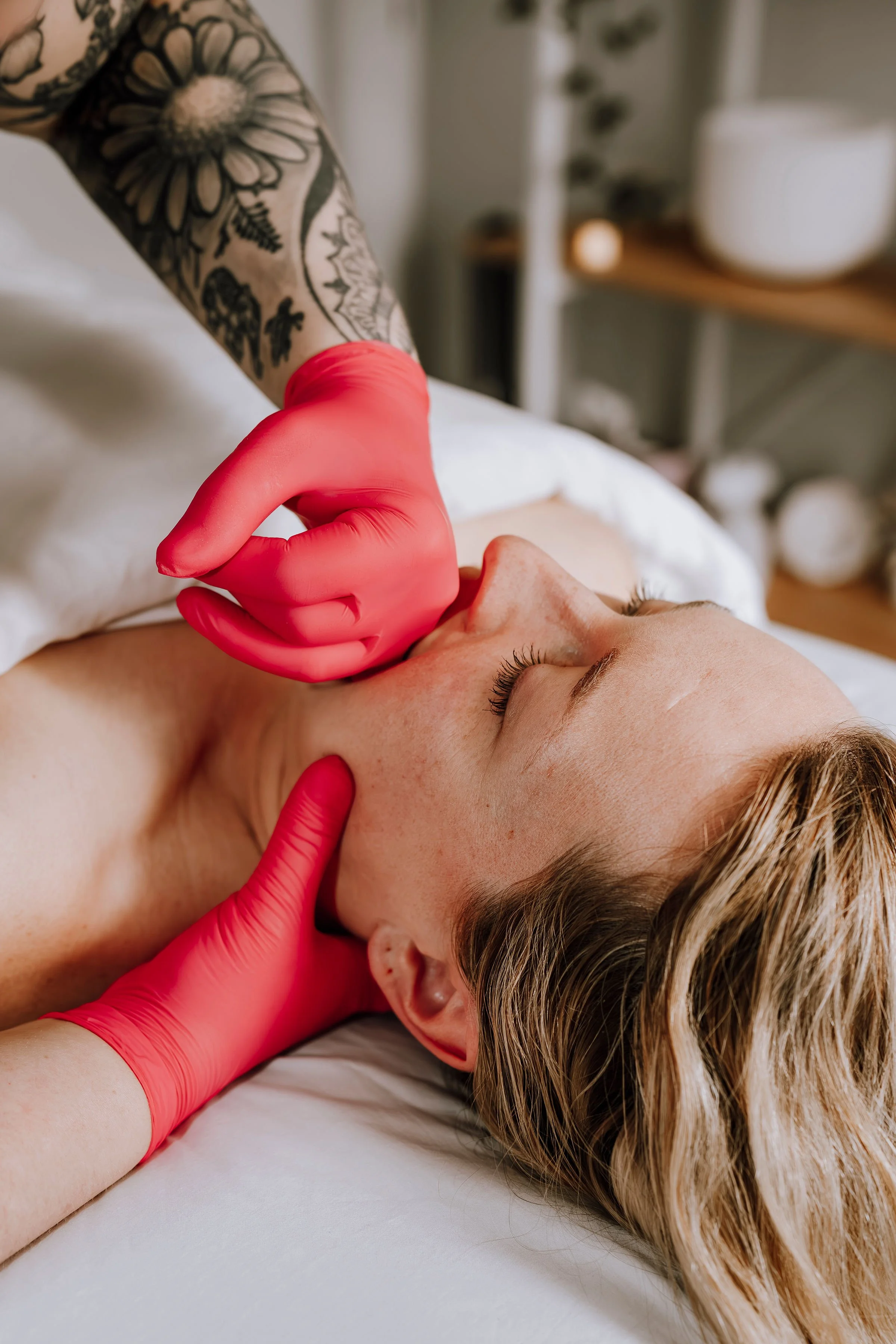A healthcare professional wearing pink gloves examines a woman's neck and jawline while she lies on a treatment bed with her eyes closed.