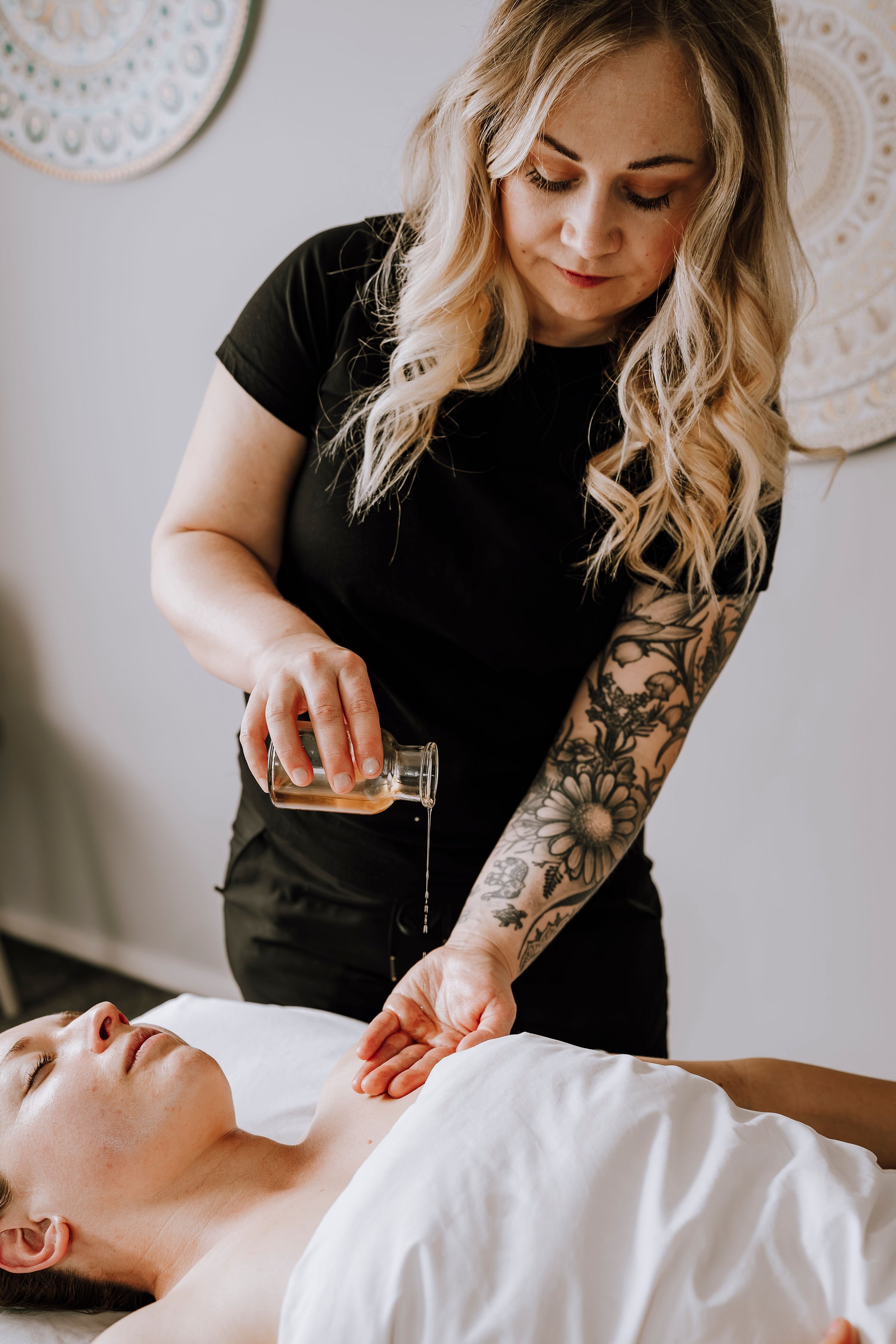 A woman with long blonde curls and tattoos on her arm is pouring oil on a man's arm while he lies on a massage table with his eyes closed.