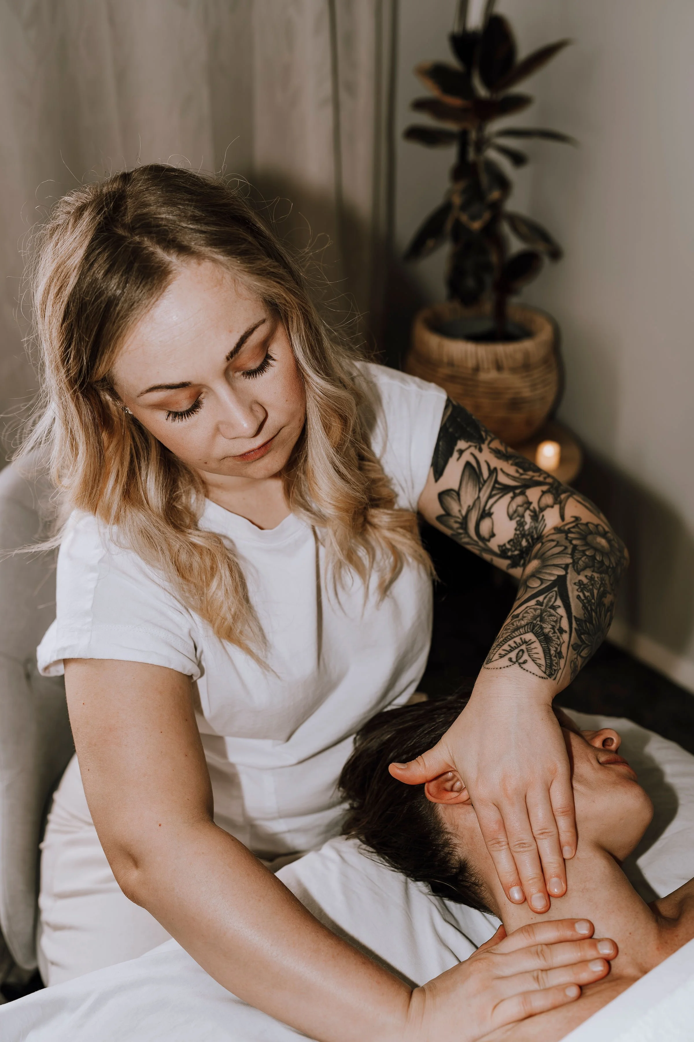 A woman with blonde hair and tattoos on her arm gives a massage to a person lying down with their head turned to the side.