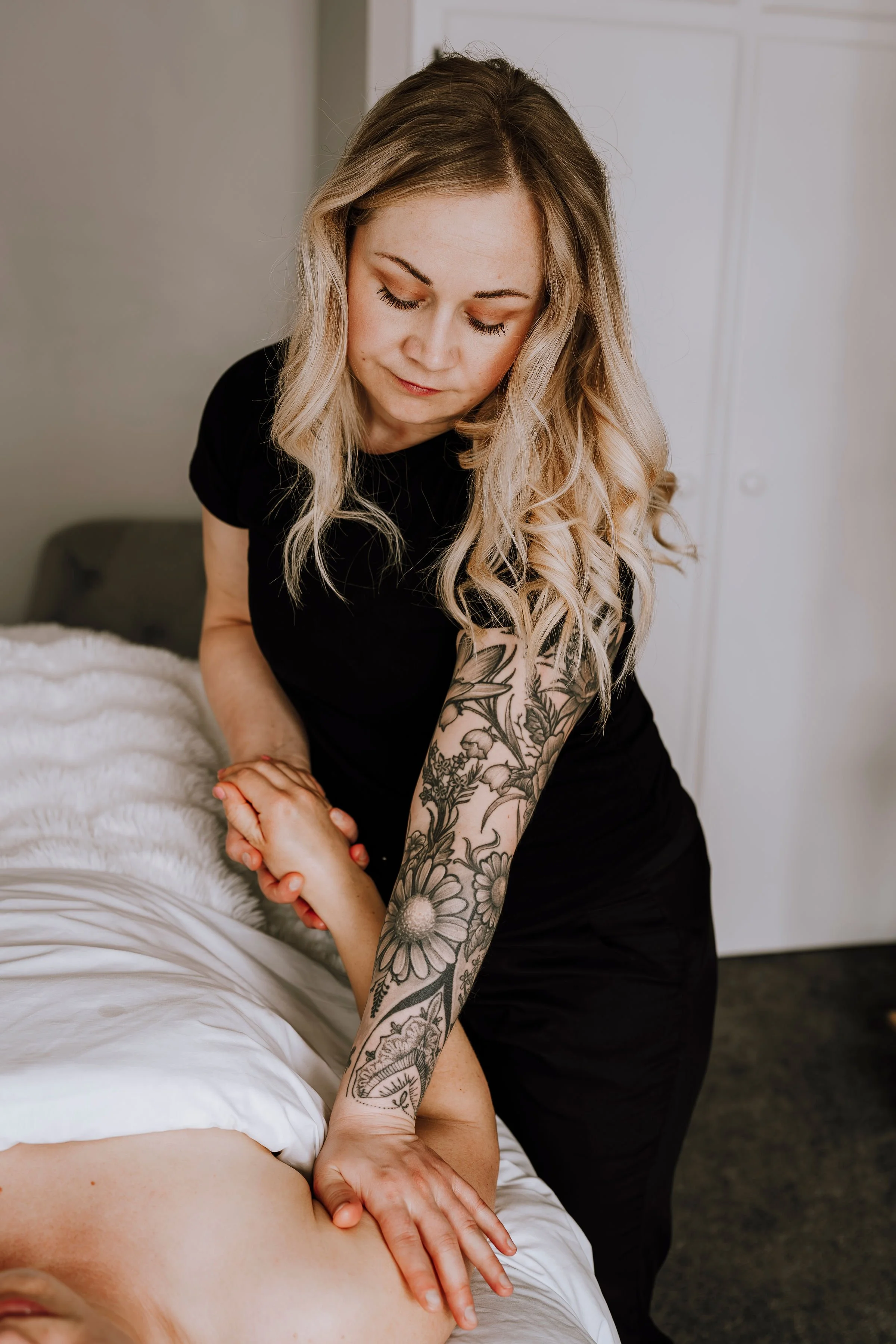 A woman with long blonde hair and a floral tattoo on her arm is giving a massage on a person's back in a room with neutral-colored walls and bedding.