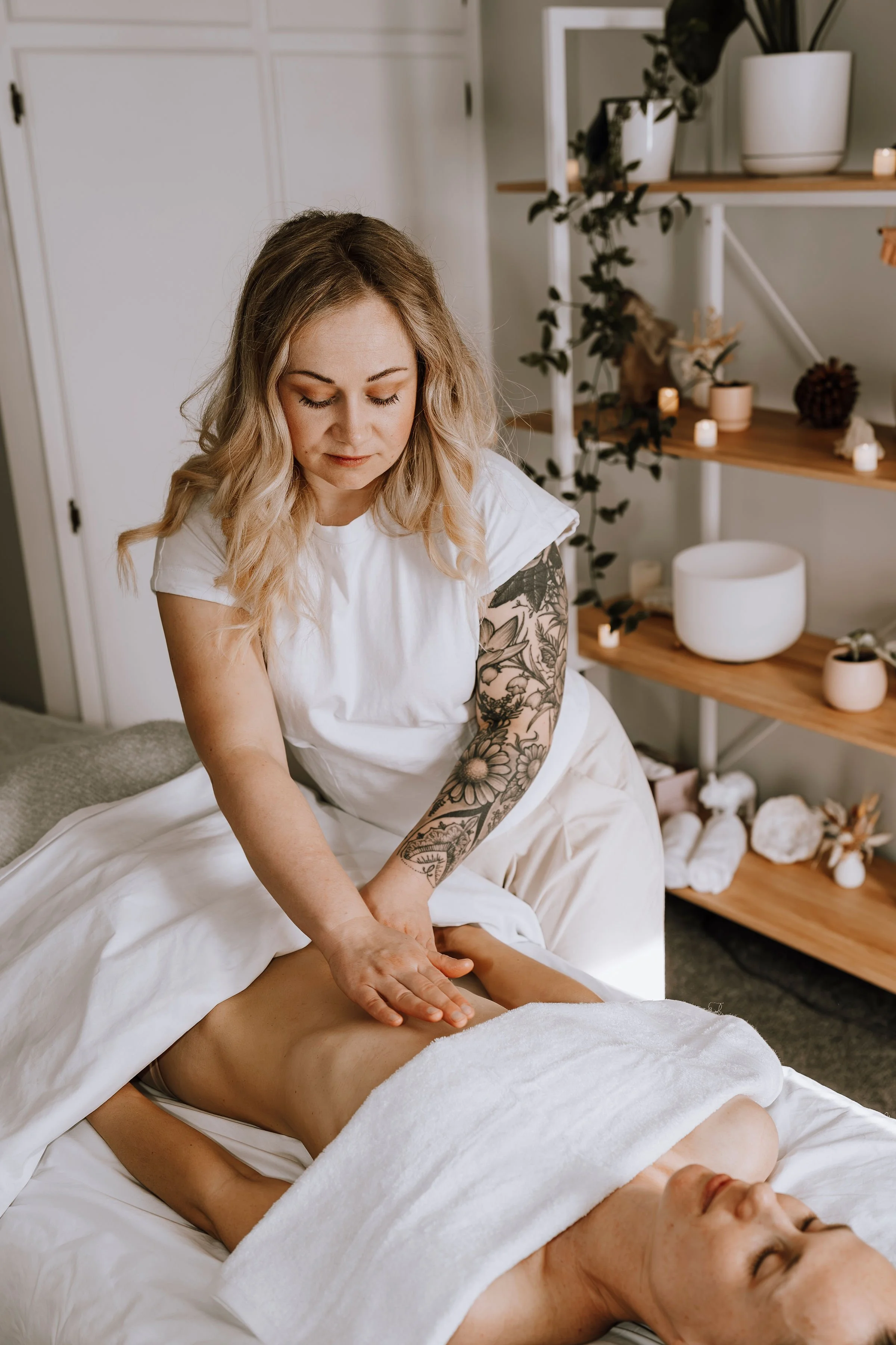 Woman giving a massage to a person lying on a massage table in a room with shelves and candles.