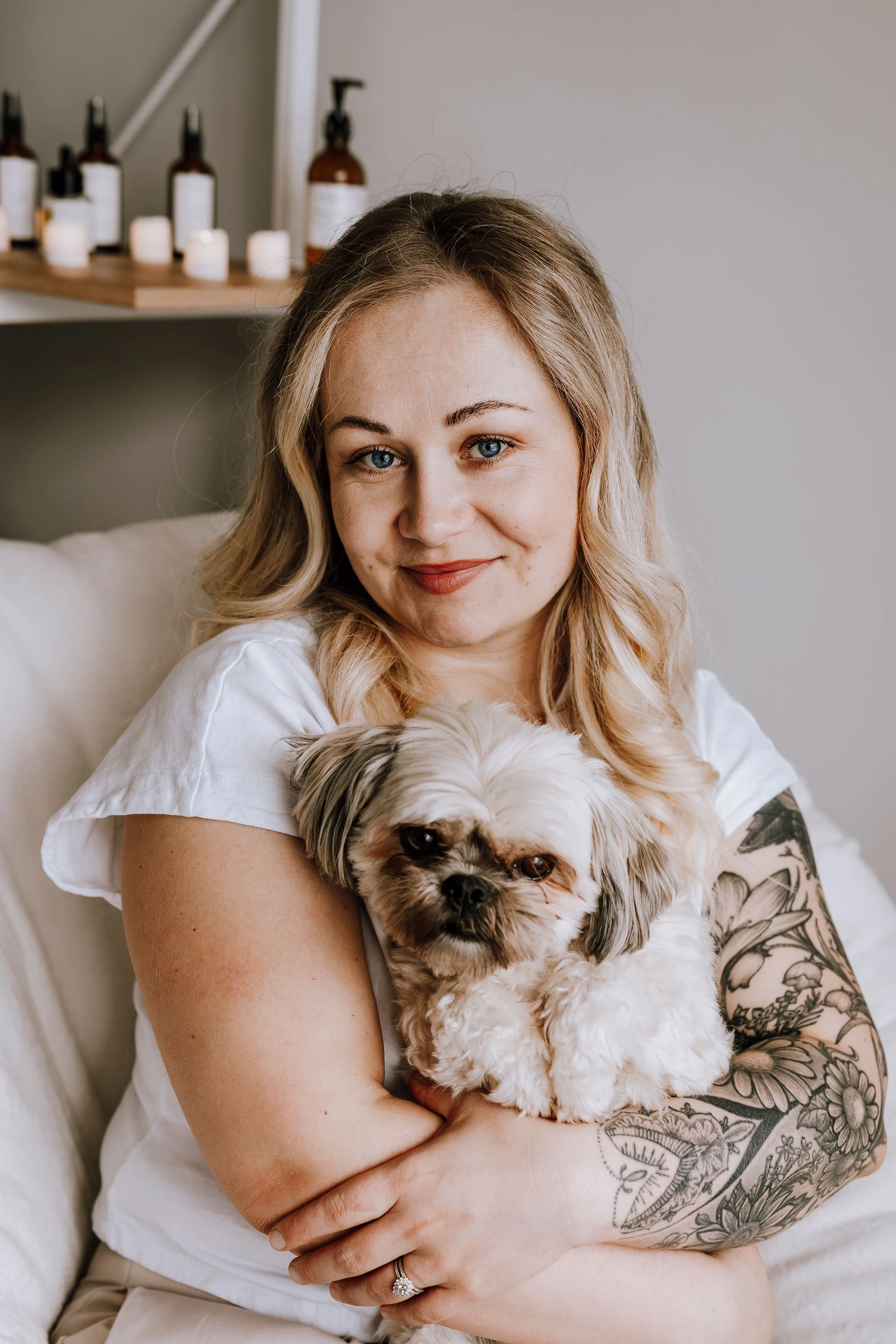 A woman with light blonde hair and a tattooed arm holds a small, fluffy dog with cream and gray fur, sitting on a white couch.