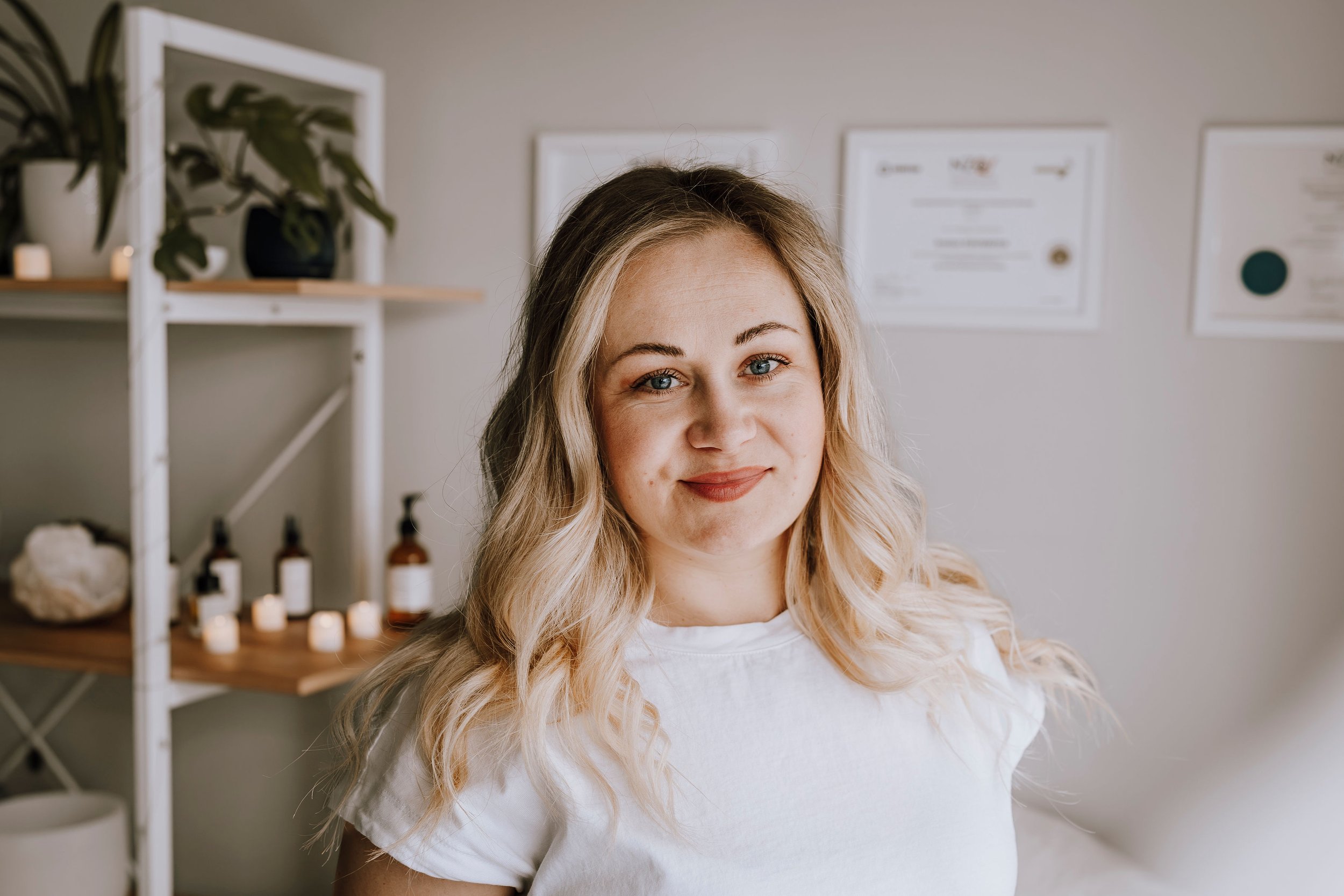 A young woman with blonde, wavy hair smiling at the camera in a wellness or spa setting.