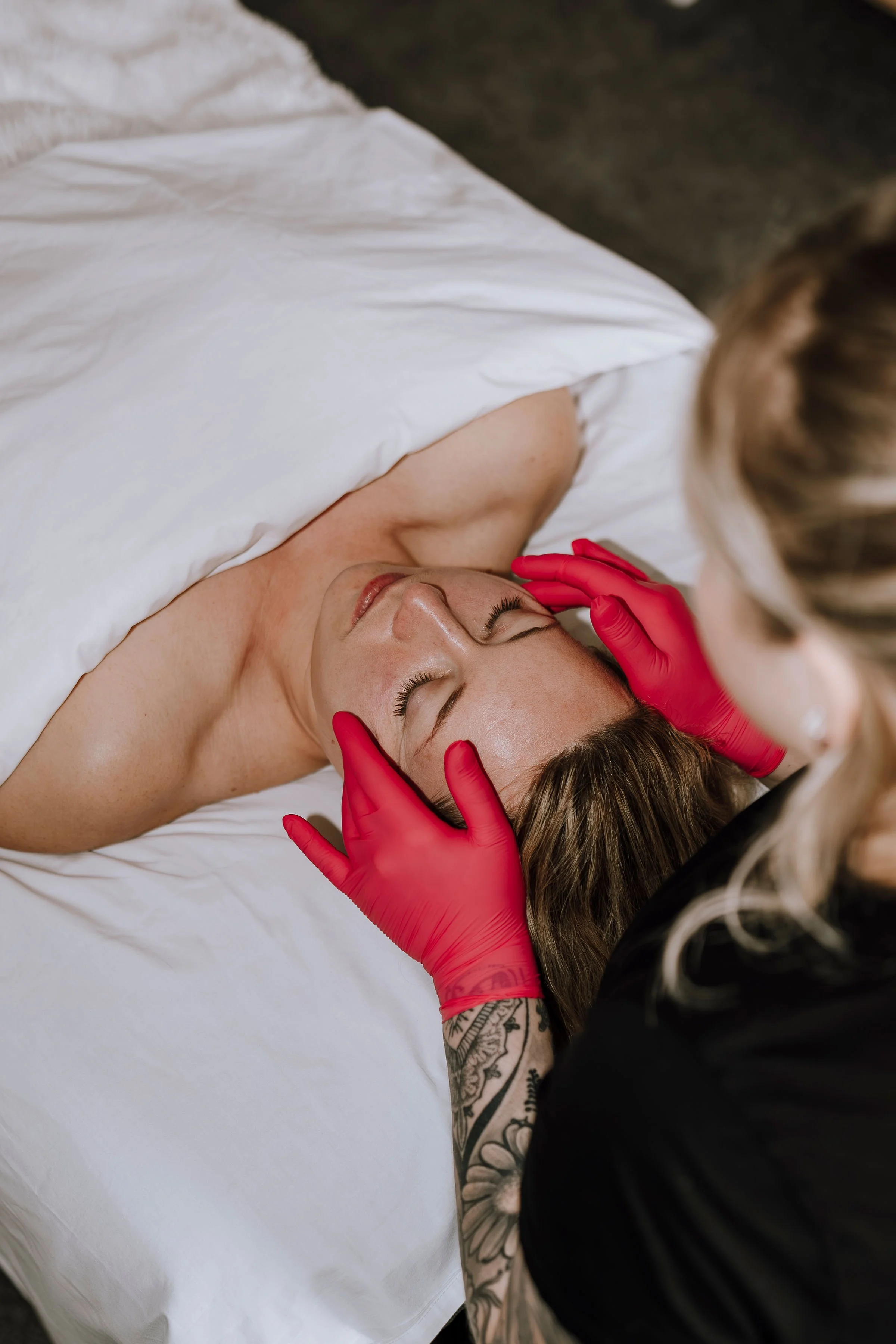 A woman receiving a facial treatment while lying on a bed covered with a white sheet, wearing a transparent facial mask, with her eyes closed, as a professional in red gloves gently presses her forehead.