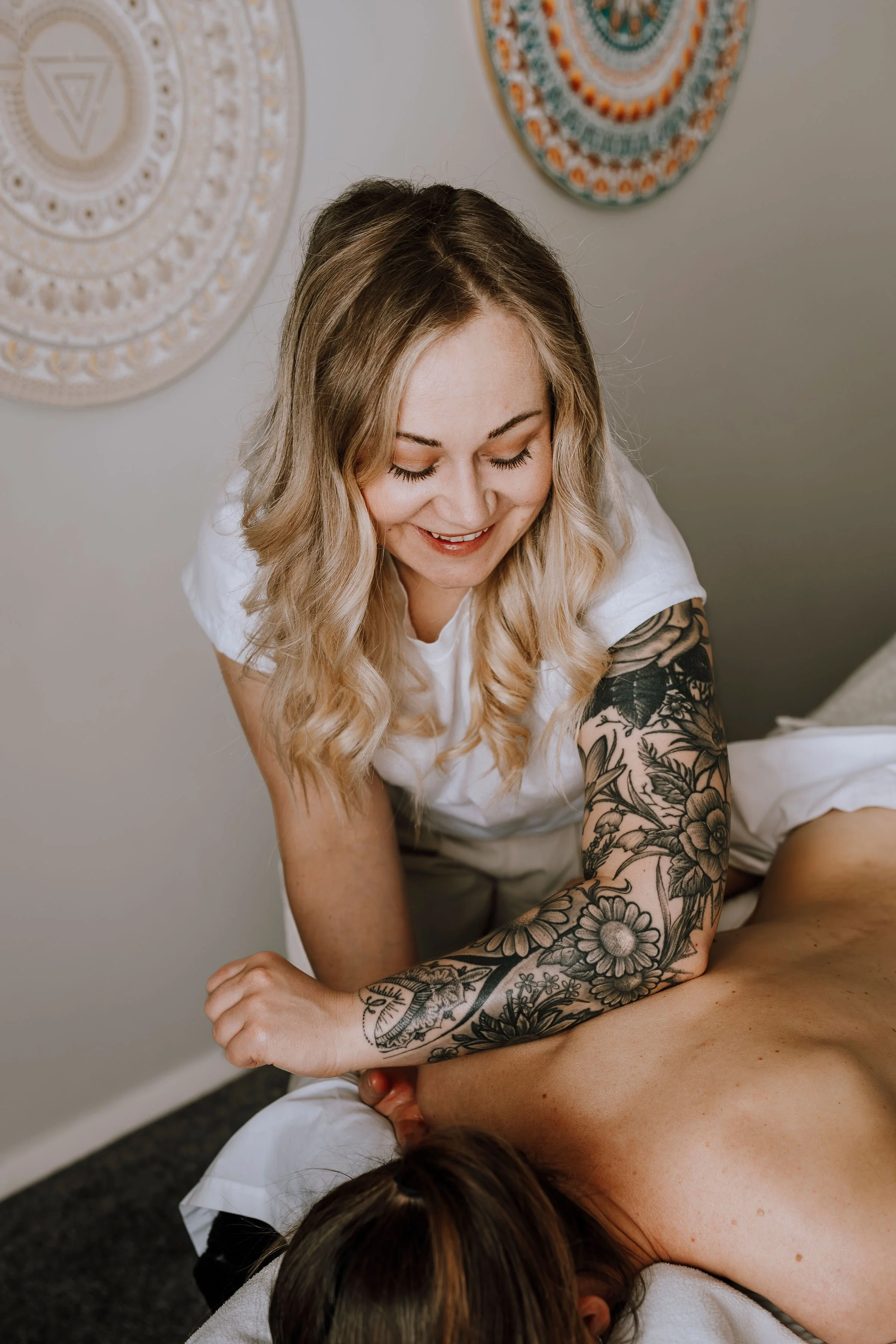 A woman with blonde hair and tattooed arm giving a massage to a person lying face down on a massage table.