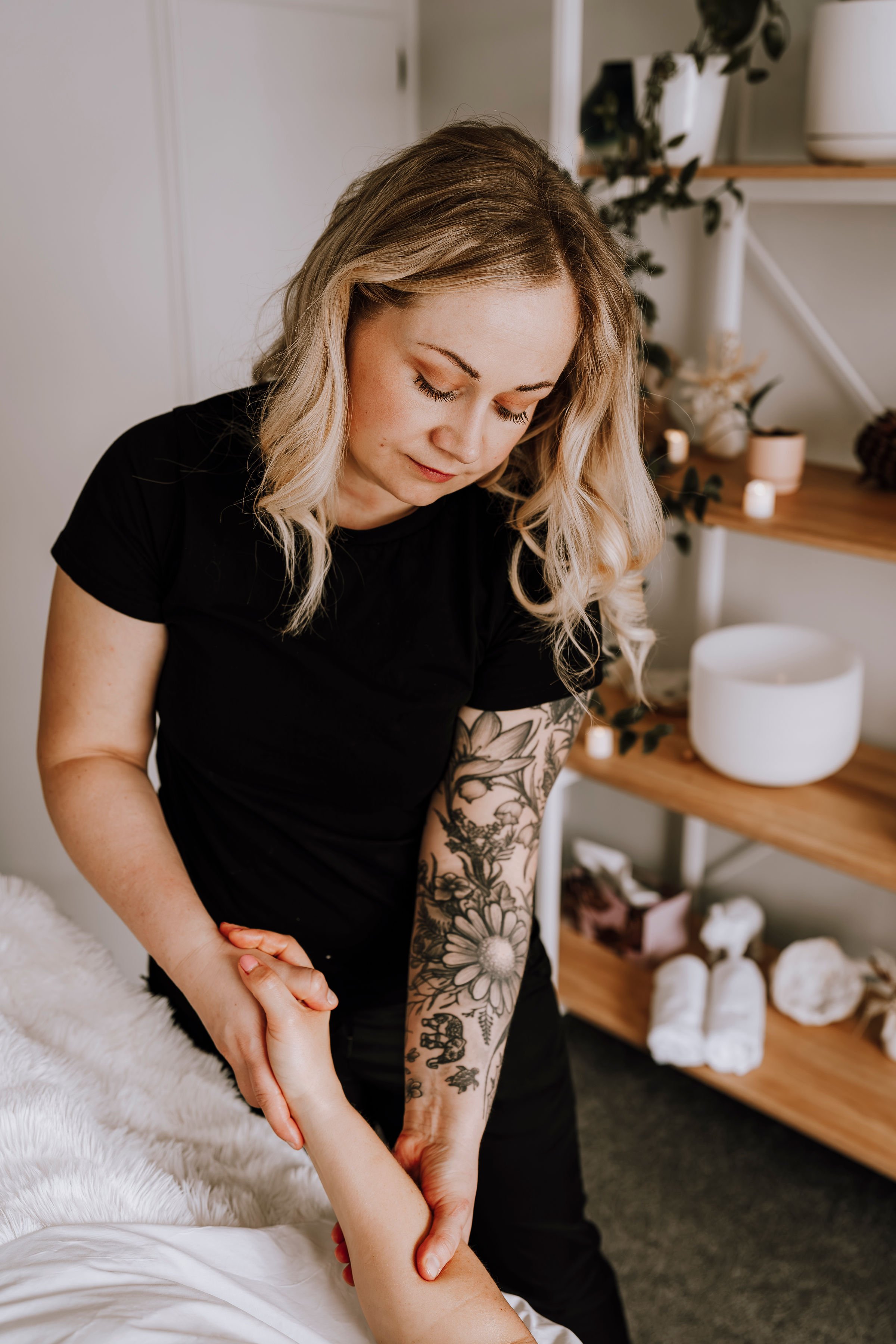 A massage therapist with a floral tattoo on her left arm giving a massage to a client in a serene, well-decorated room with wooden shelves and white candles.