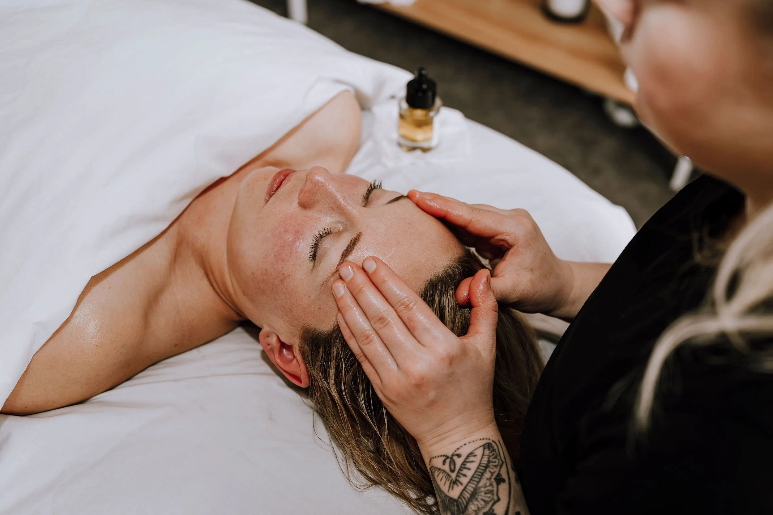 A woman receiving a facial treatment, lying on a bed with her eyes closed, as a skincare professional gently massages her forehead. A small bottle of facial oil is on the bed beside her.