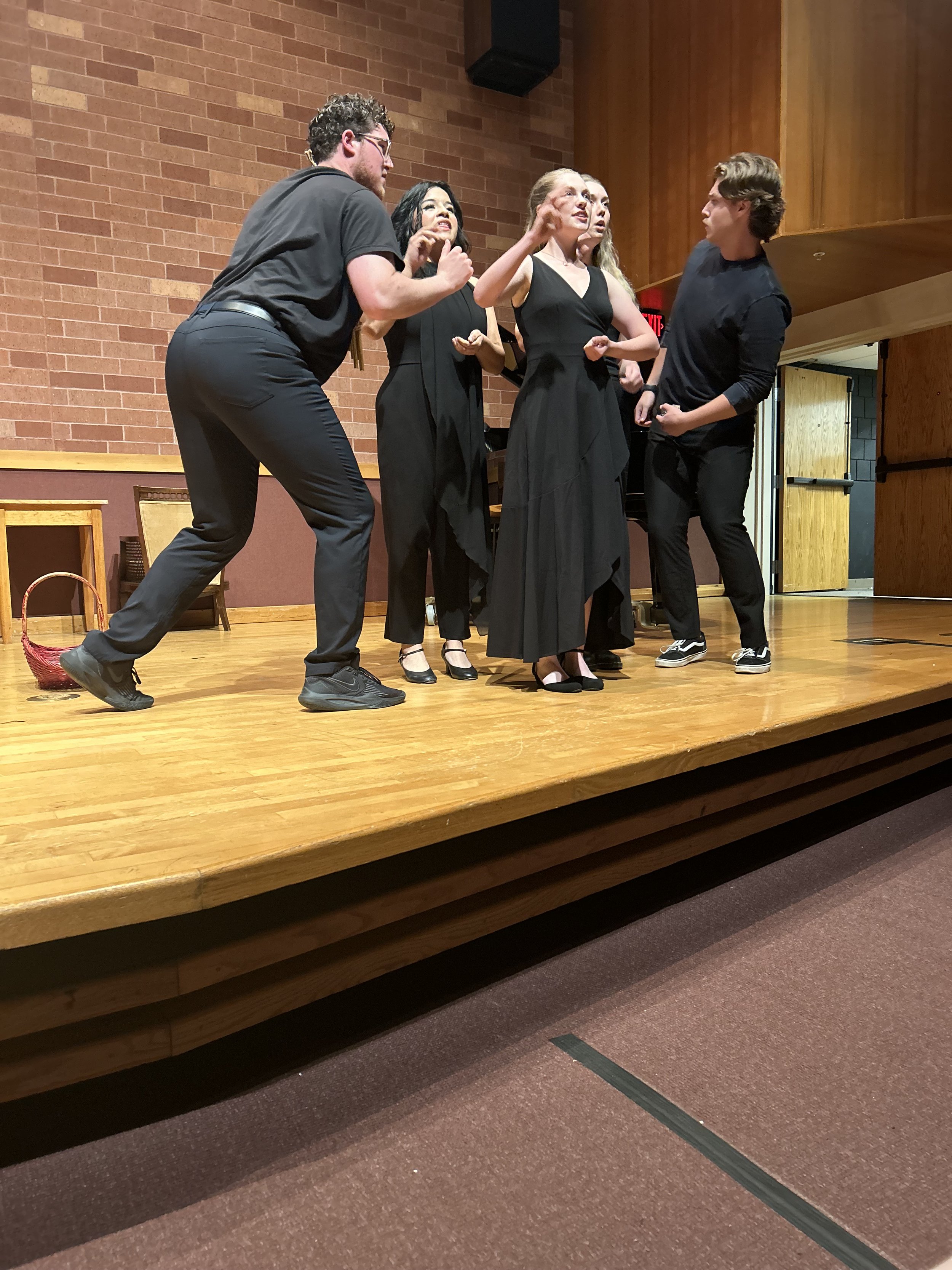 Group of five actors engaged in a theatrical scene on a stage with a brick wall background, two women dressed in black, three men in dark clothing, one man appears to be arguing or accusing, and the women seem to be responding.