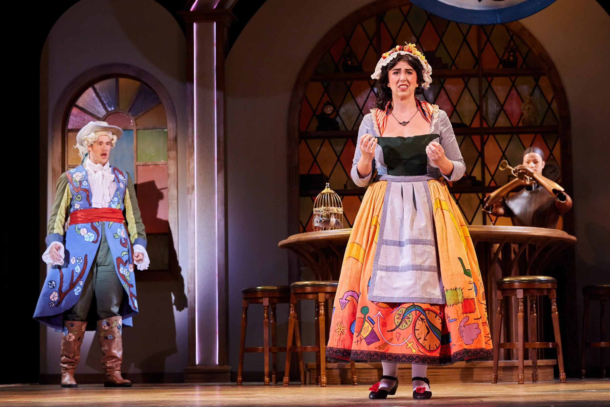 Three actors on stage performing in a play, dressed in colorful, period costumes. The woman in the center wears a long skirt with bright patterns and an apron, looking distressed. A man on the left wears a blue coat with embroidered floral designs an