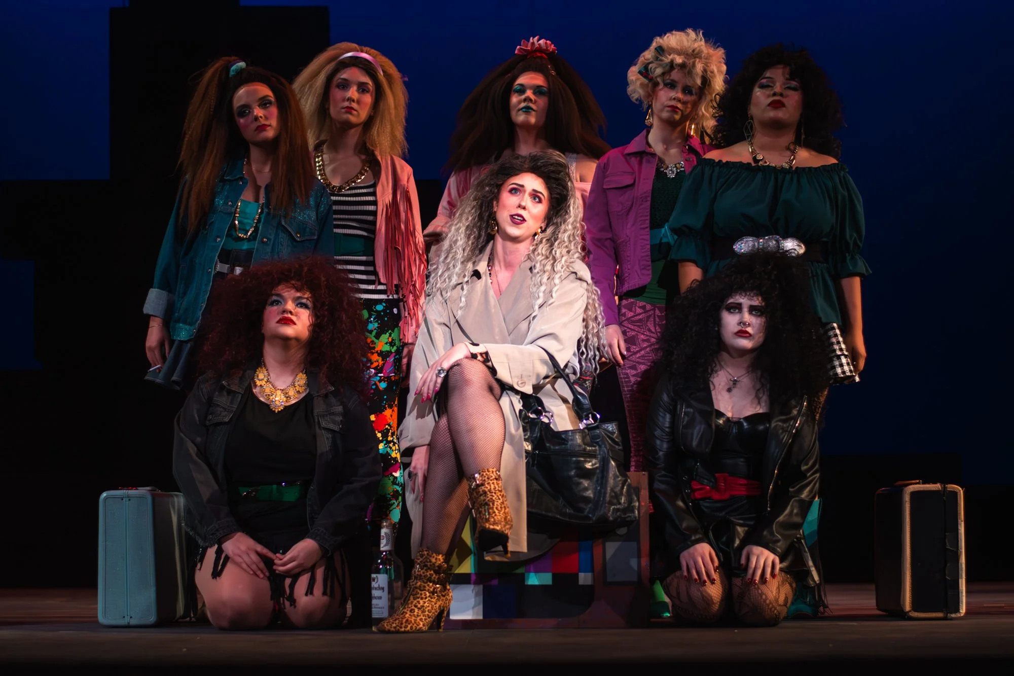 Group of women with vibrant 80s-style fashion and big hair, posing on a stage with a dark background.