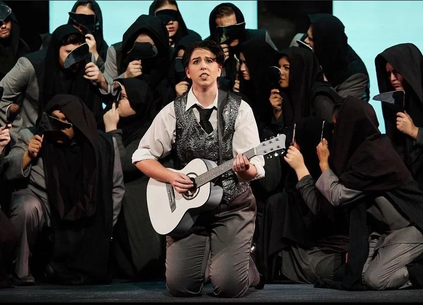 A woman singing with a guitar, kneeling on stage surrounded by people wearing black hoods and masks.