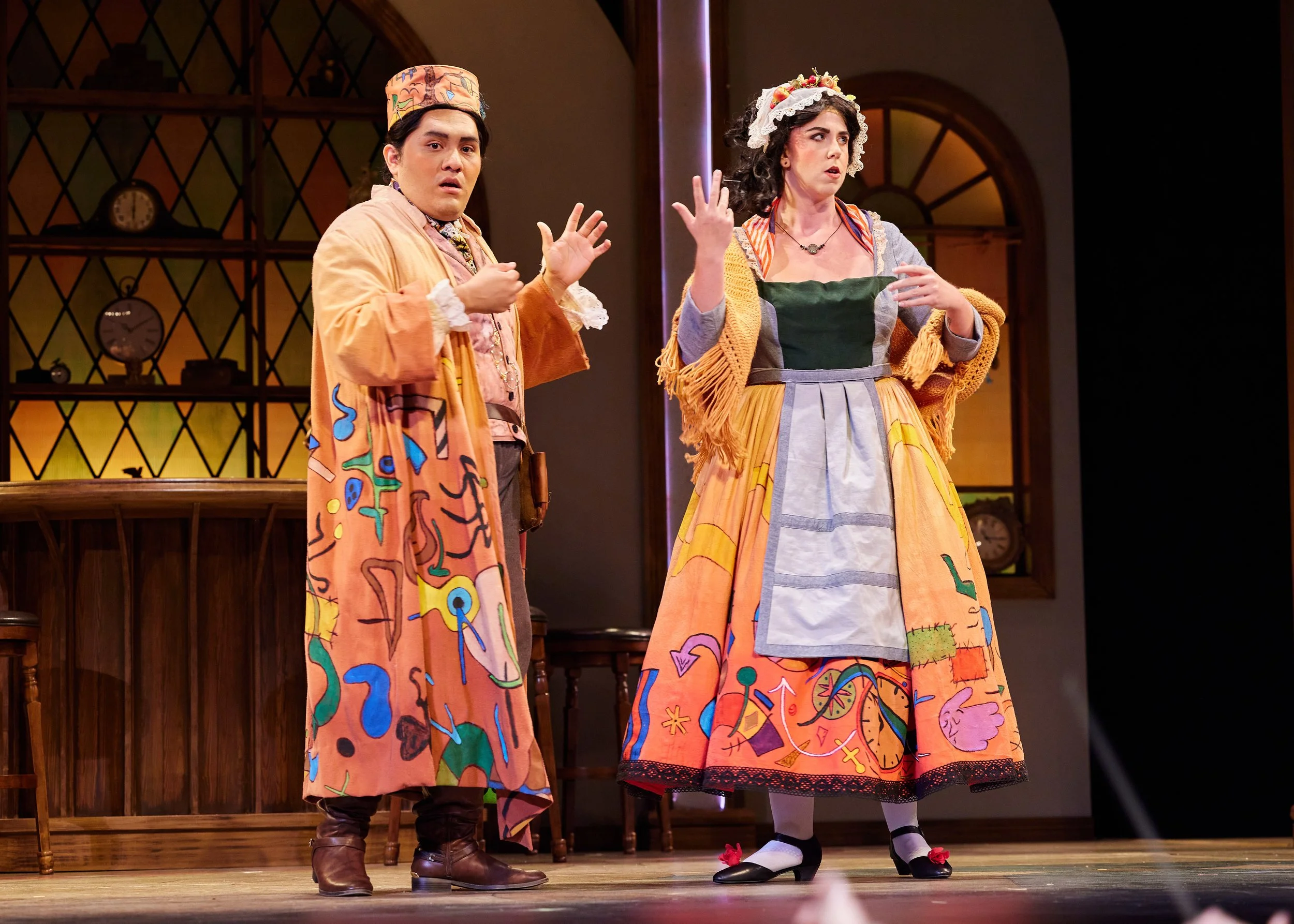 Two actors in colorful, elaborate costumes perform on stage against a wooden backdrop.