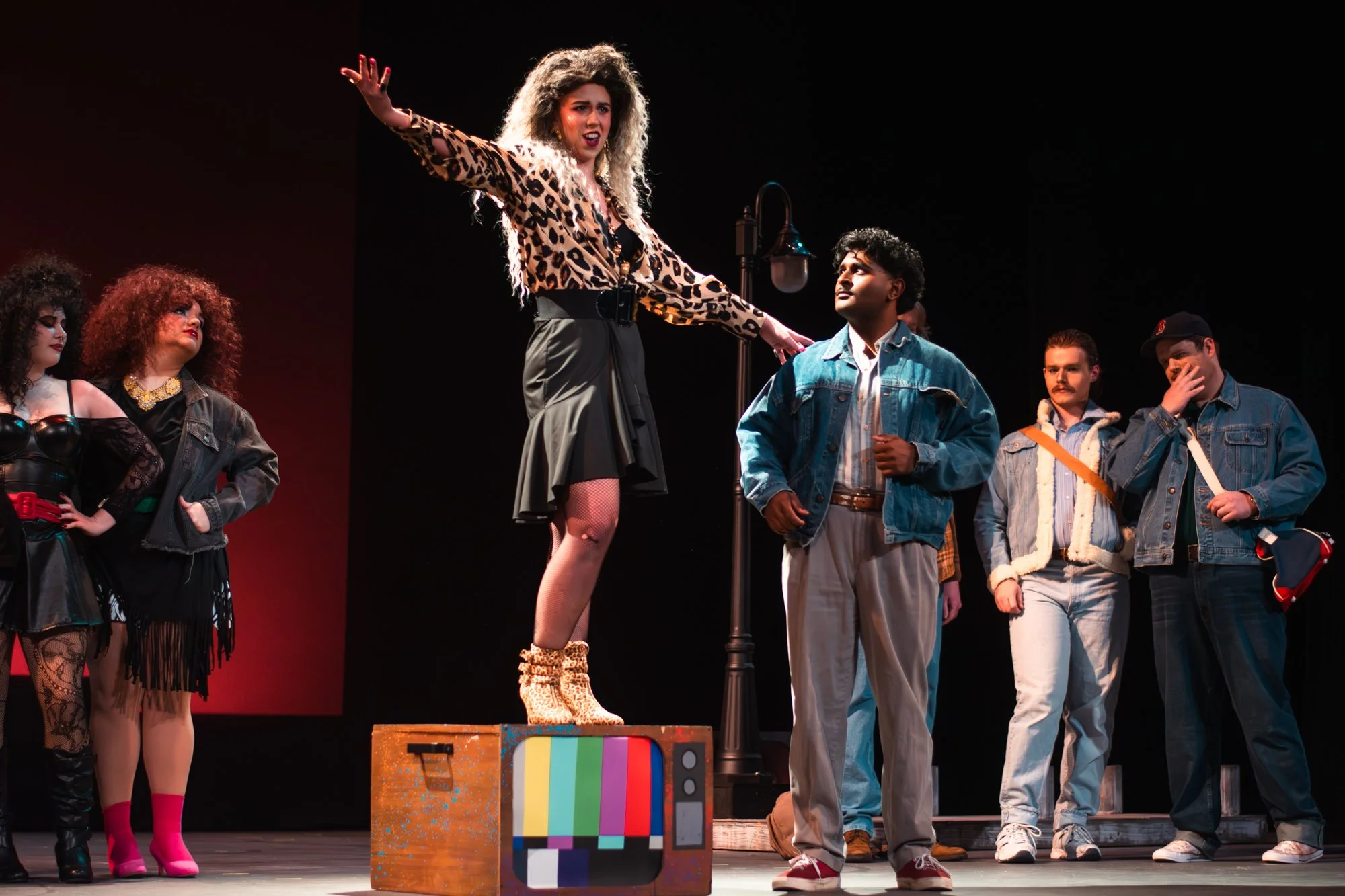 People performing on stage in a theatrical play, with a woman standing on a box and others standing nearby, in casual and theatrical costumes.