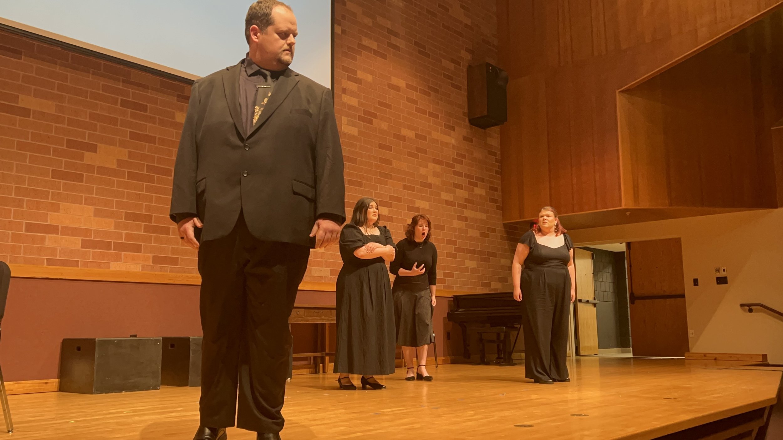 Four people standing on a wooden stage in a large room with brick walls, possibly during a theatrical or musical performance. The man in the foreground wears a black suit, and three women in the background are dressed in black, with two of them displ