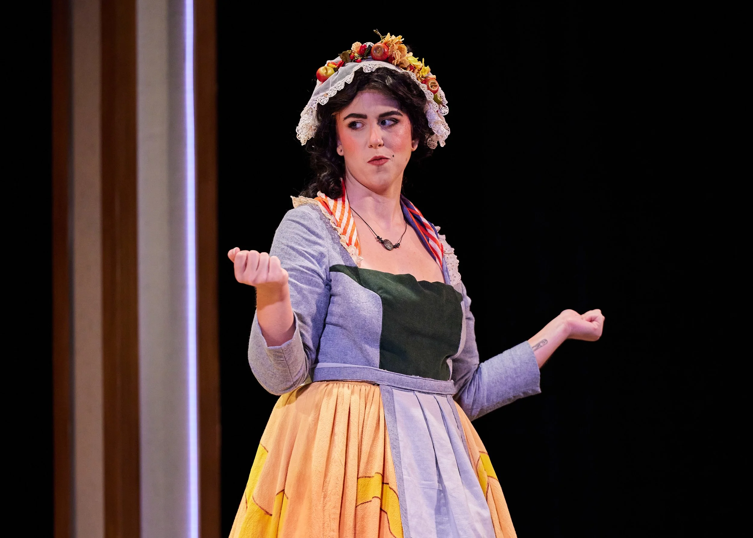 Women dressed in traditional European folk costume, wearing a floral headpiece, gray blouse, and colorful skirt, standing on stage with an expressive facial expression and gesturing with her hands.